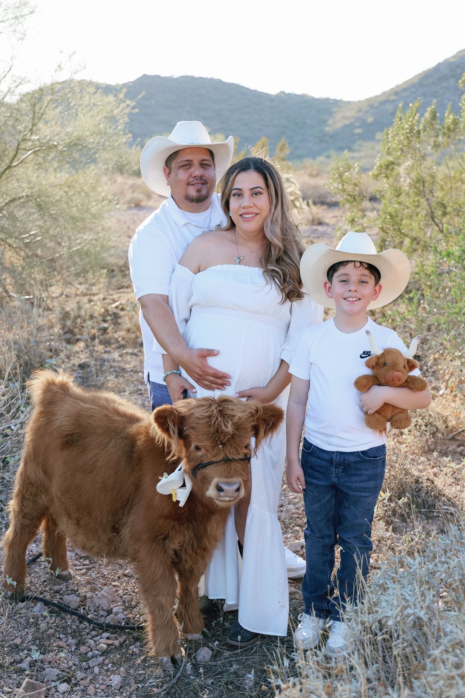 Pregnant woman in white dress with husband and son in cowboy hats posing with a fluffy Highland calf outdoors.
