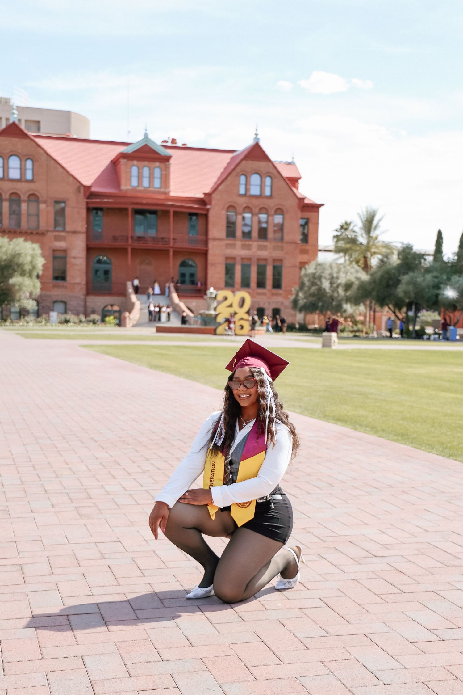 A smiling graduate in cap and gown kneels on a brick path in front of a red brick university building.