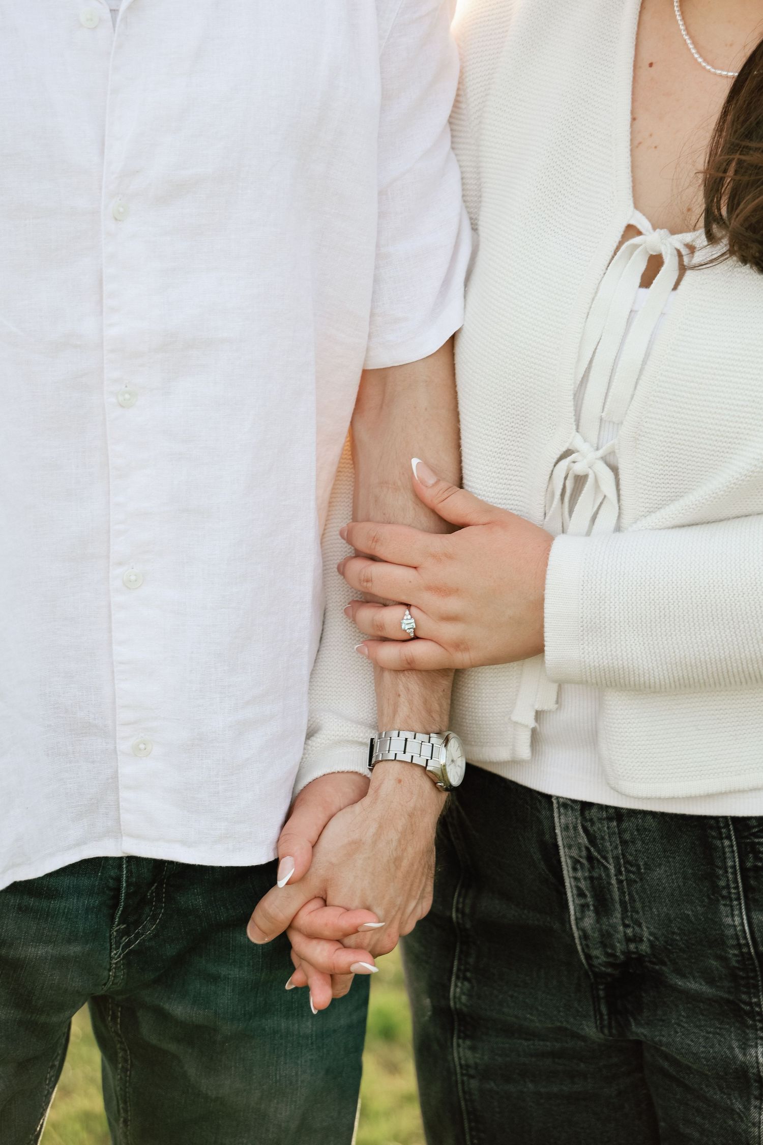 Couple holding hands showing engagement ring and watch, wearing white tops and dark jeans outdoors.