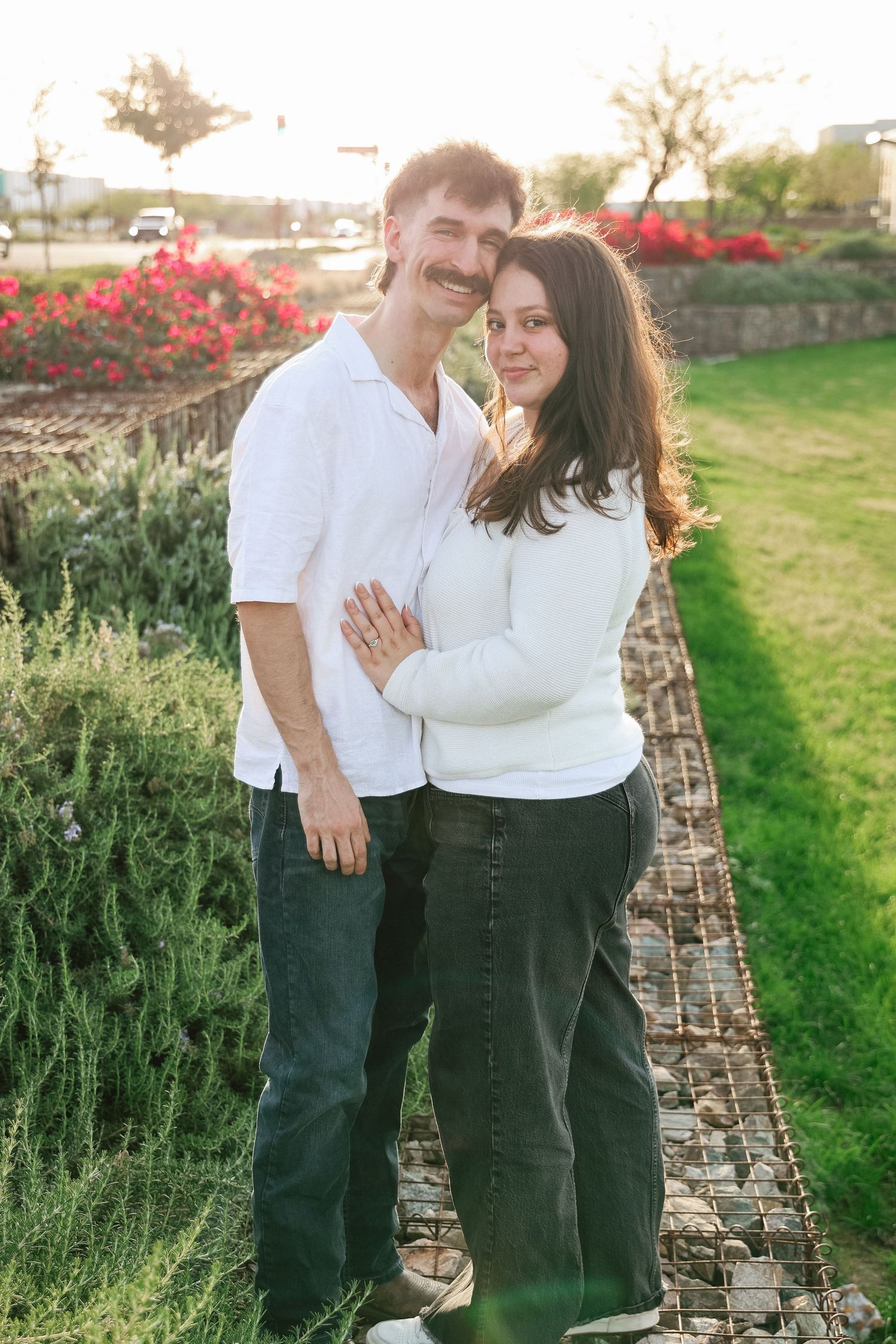 A smiling couple embracing outdoors near a garden with red flowers and green grass during golden hour.