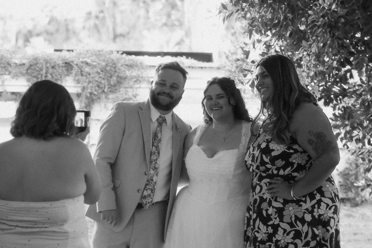Black and white photo of smiling bride and groom posing with a guest at their outdoor wedding.