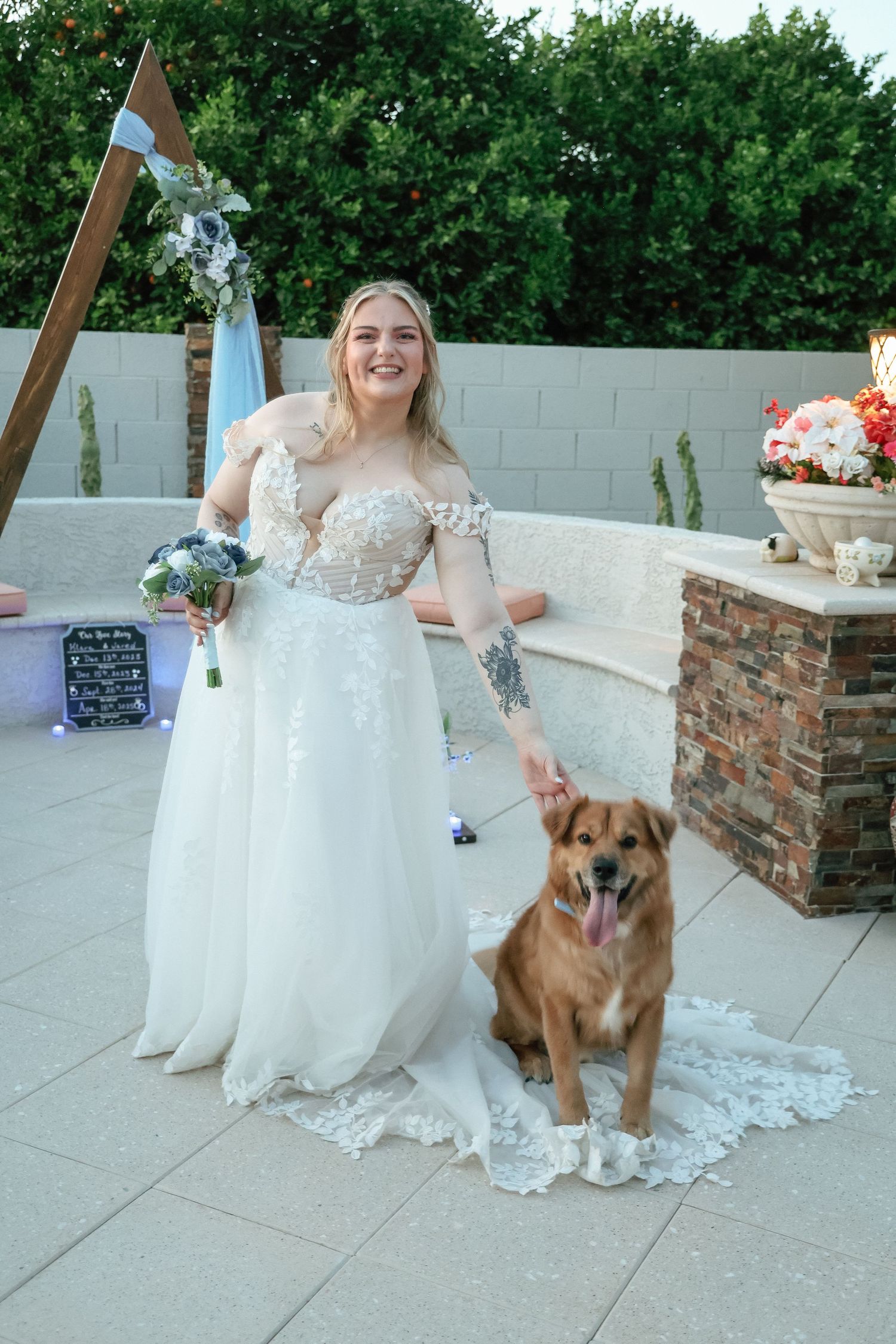 Smiling bride in lace wedding gown holding bouquet poses with golden dog near triangular wooden arch.