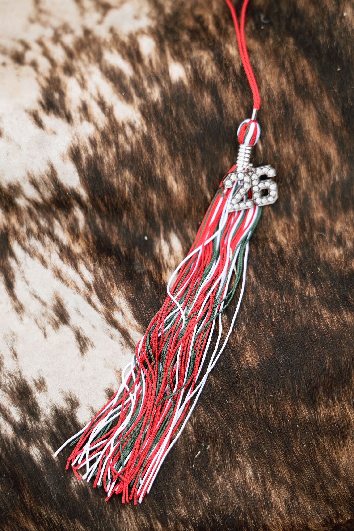 Red, white, and green graduation tassel with silver 26 charm on cowhide background.