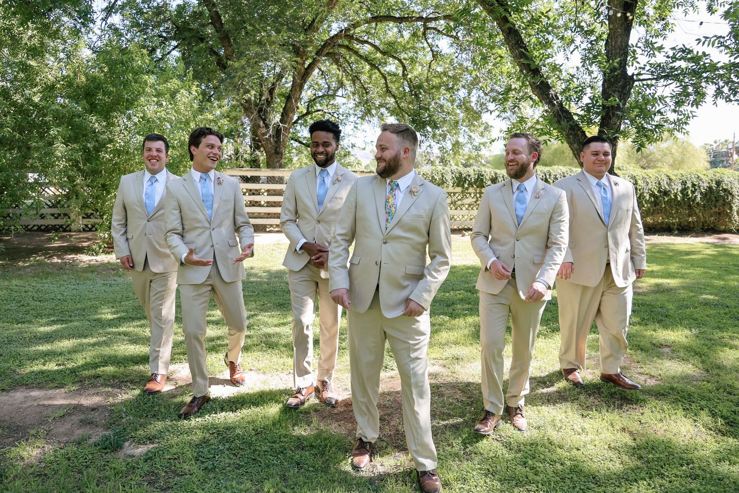 Six groomsmen in matching tan suits with floral ties pose outdoors on green grass beneath large shade trees.