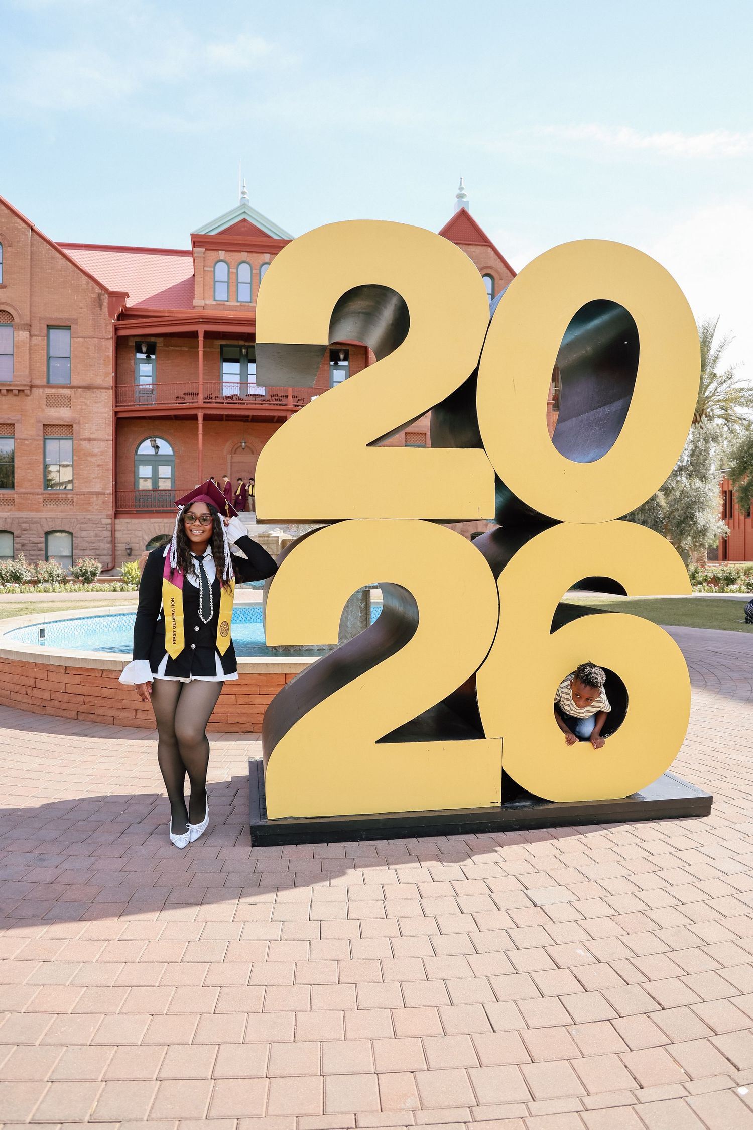 Student posing beside a large gold 2026 sign on a brick plaza outside a red brick university building.