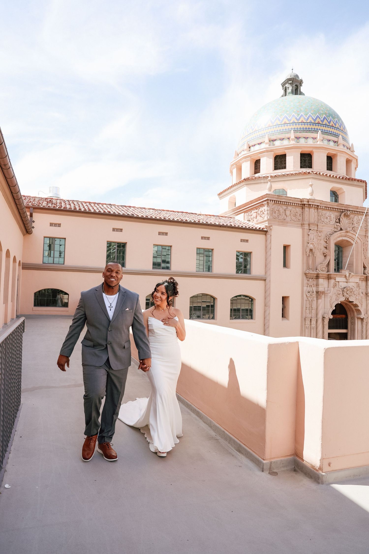 Bride and groom walking hand in hand on a rooftop terrace with a historic domed building backdrop on their wedding day.
