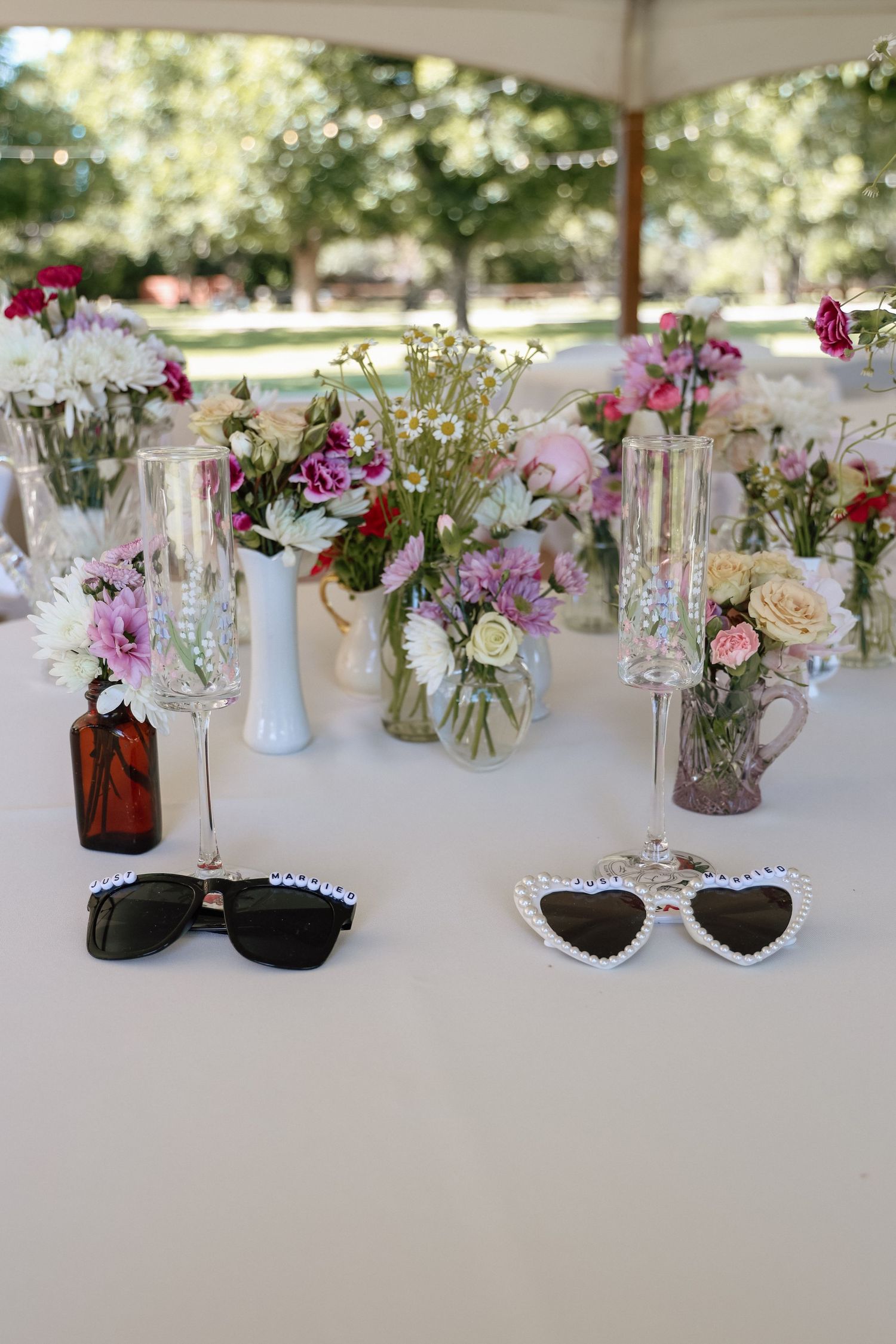 Elegant outdoor wedding reception table with floral centerpieces, champagne flutes, and novelty sunglasses on white linen.