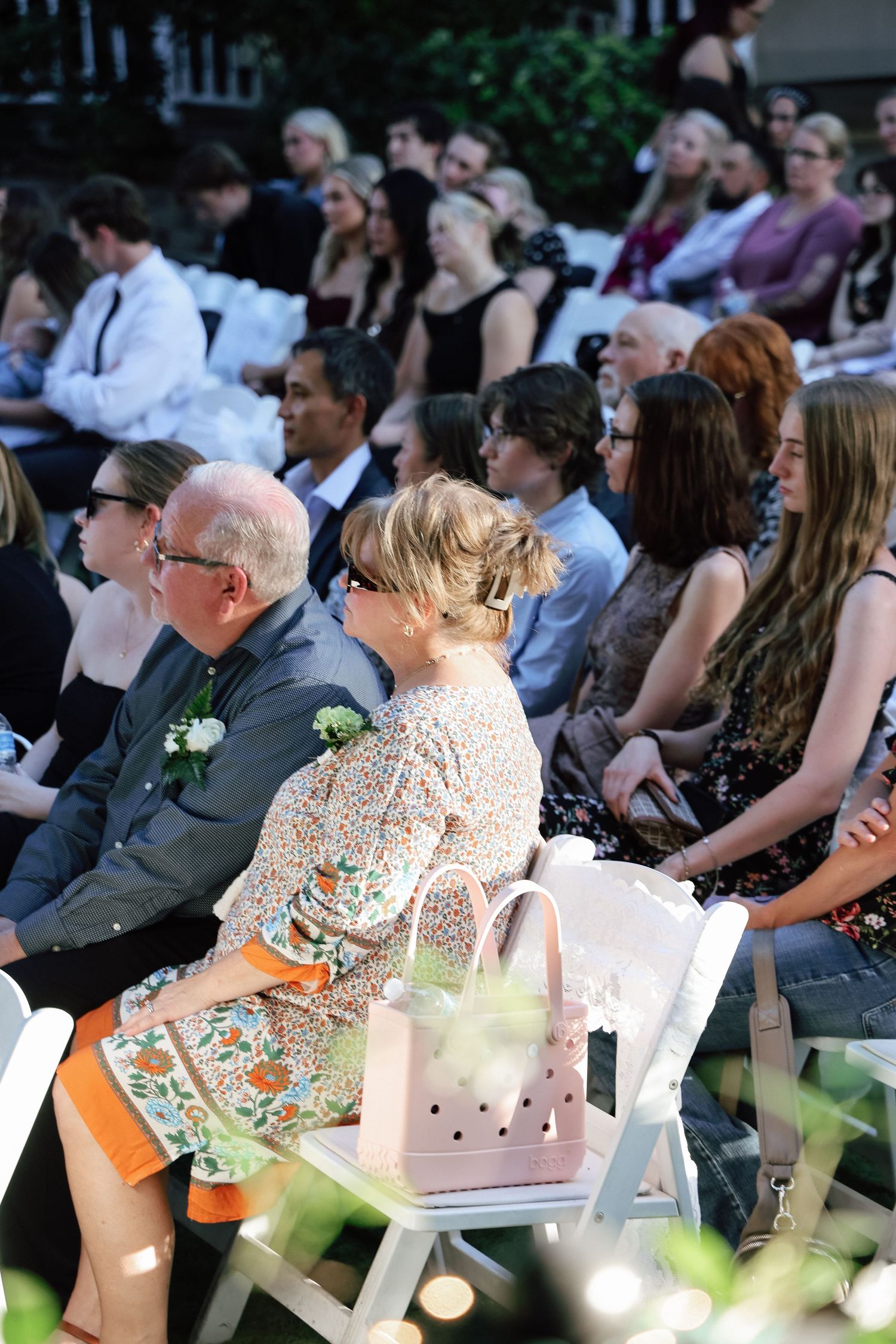 Guests seated in white chairs at an outdoor wedding ceremony, with a pink handbag resting on a chair in the foreground.