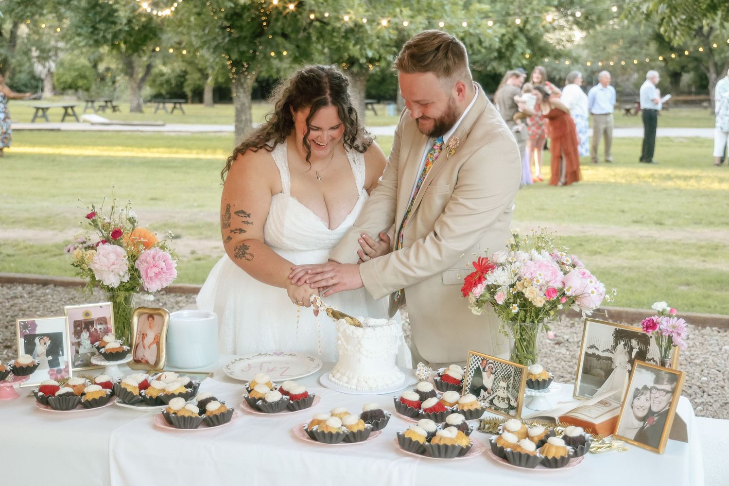 Bride and groom cutting wedding cake at outdoor reception table with cupcakes, florals, and string lights.