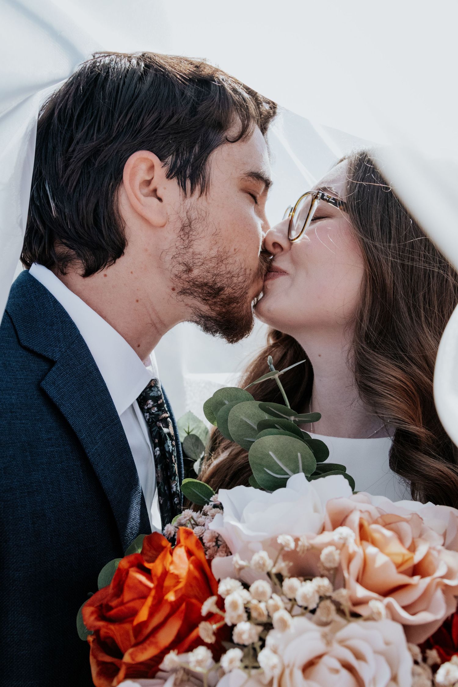 Close-up wedding photo showing a tender kiss with coral and peach roses in the foreground.