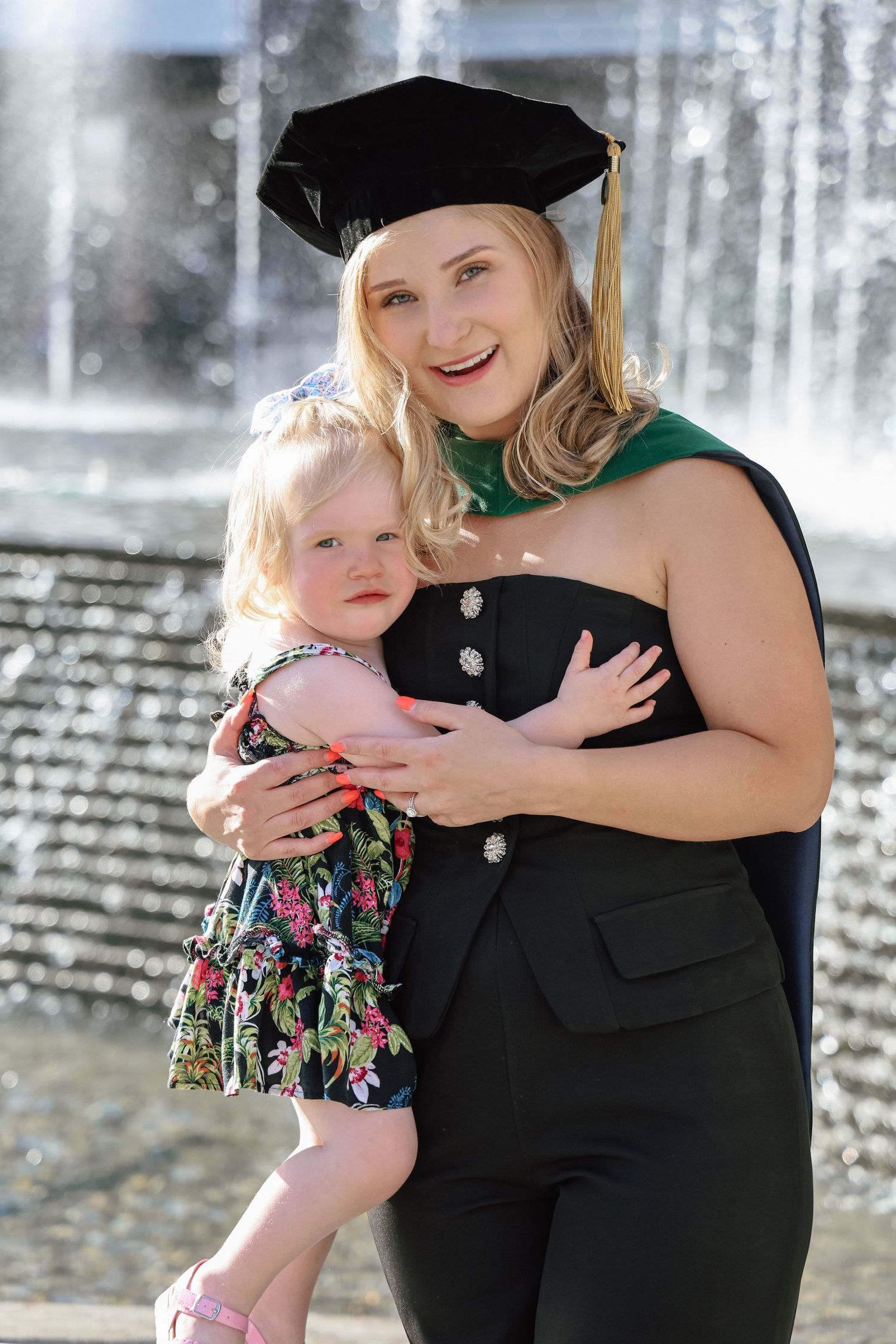 Graduate in cap and gown holding a young toddler girl in front of a fountain.