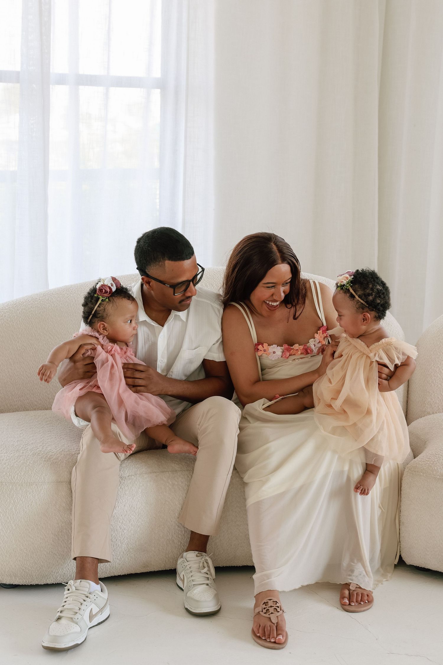 Happy family of four sitting on a white couch, parents smiling at two young daughters in pink and beige dresses.