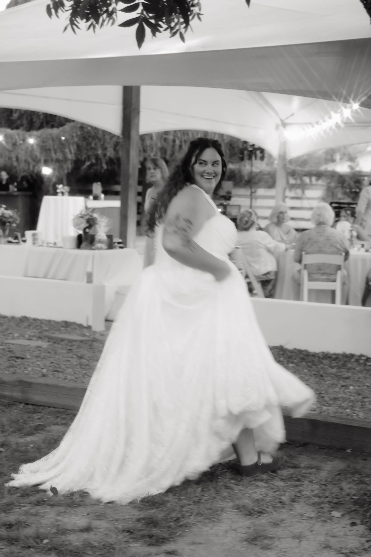 Black and white photo of bride twirling in full white gown at outdoor wedding reception under white tent.