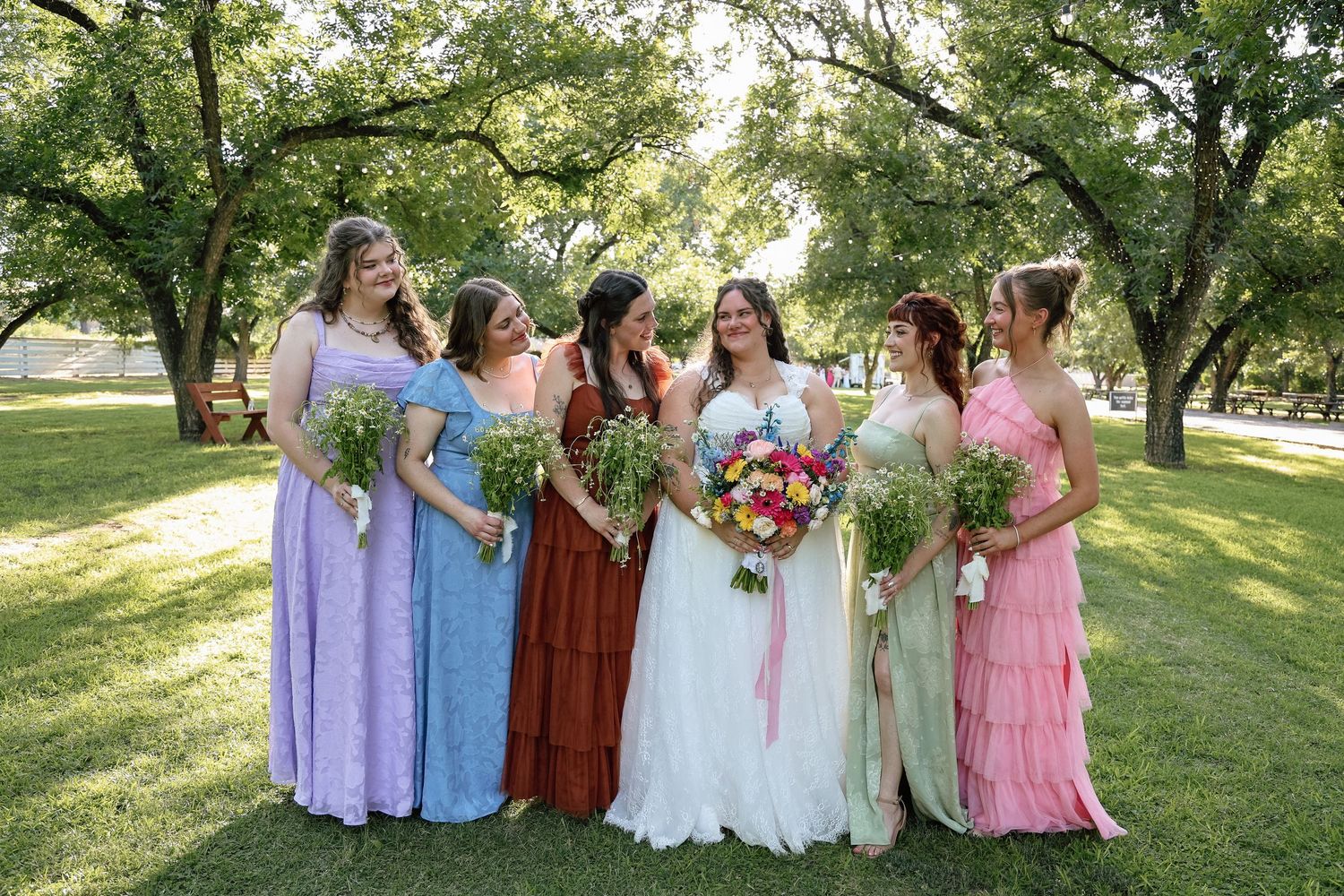 Bride in white gown with colorful bouquet stands with five bridesmaids in mismatched dresses outdoors.