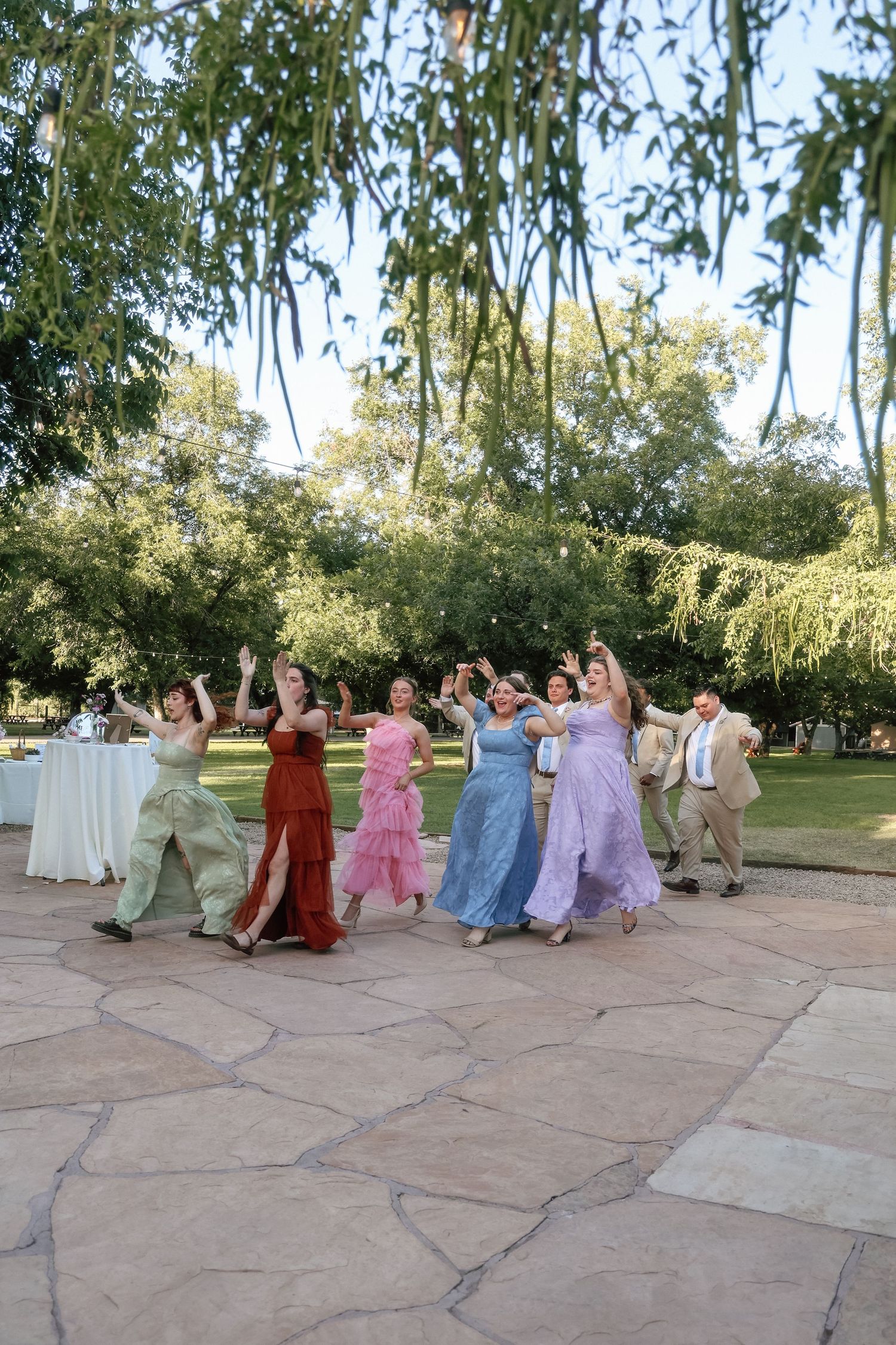 Wedding guests in colorful formal gowns dance and celebrate outdoors on a stone patio surrounded by trees.