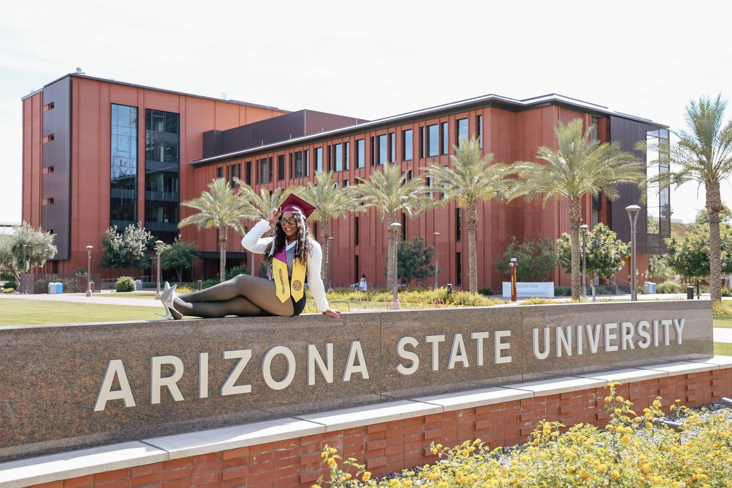 Graduate poses proudly in cap and gown at Arizona State University sign with modern campus building and palm trees.