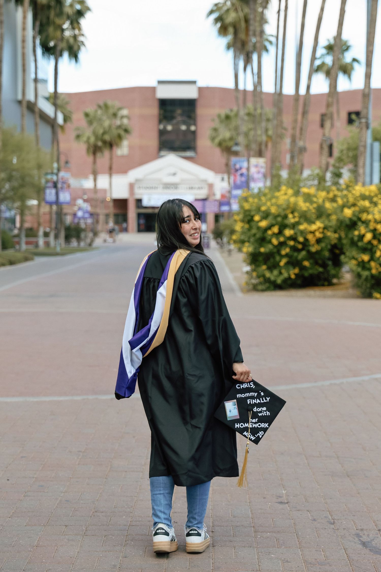 Smiling graduate in black cap and gown poses on university campus walkway lined with palm trees.