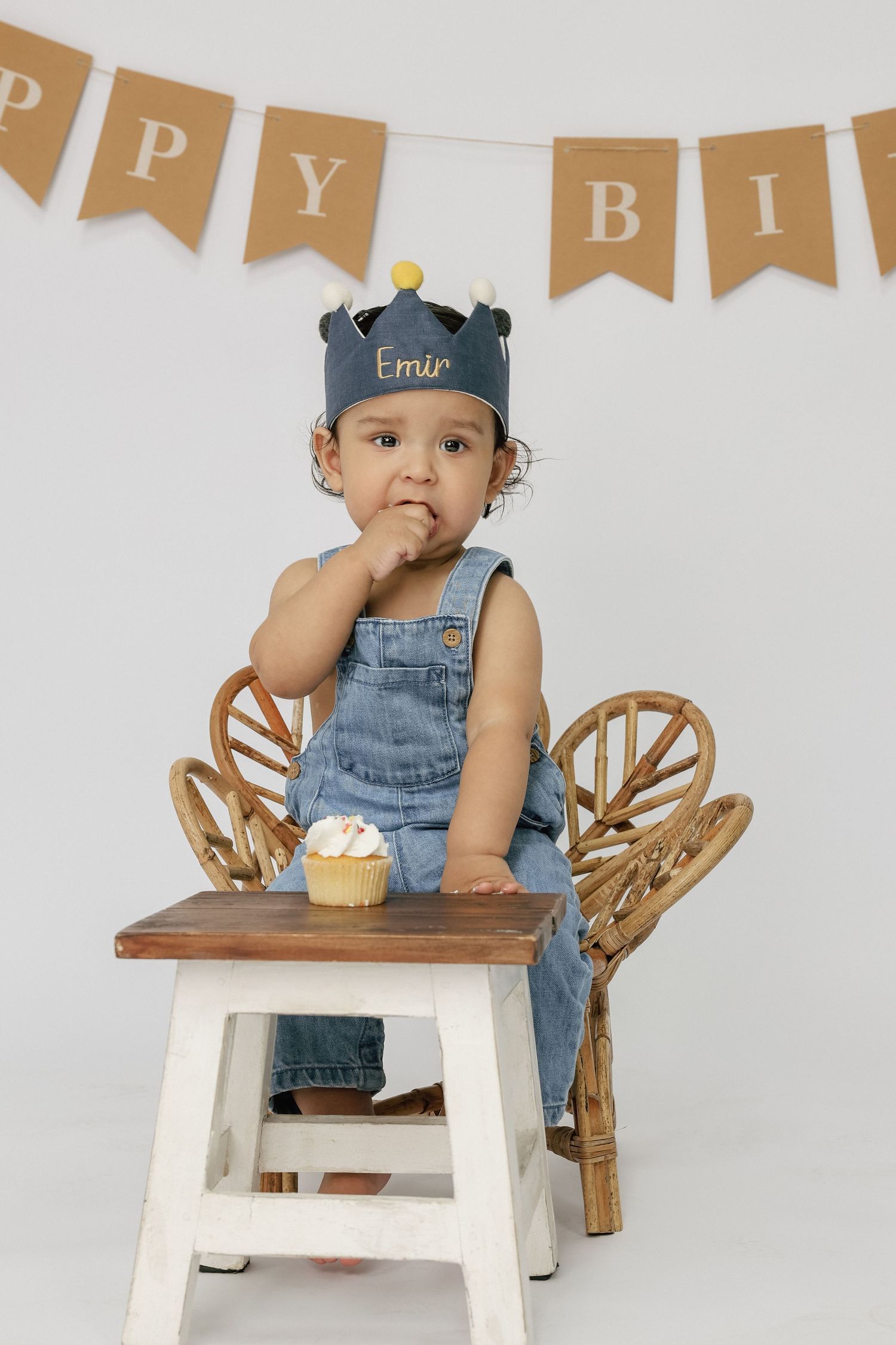 Toddler named Emir in denim overalls and birthday crown sits at small table with cupcake in front of Happy Birthday banner.