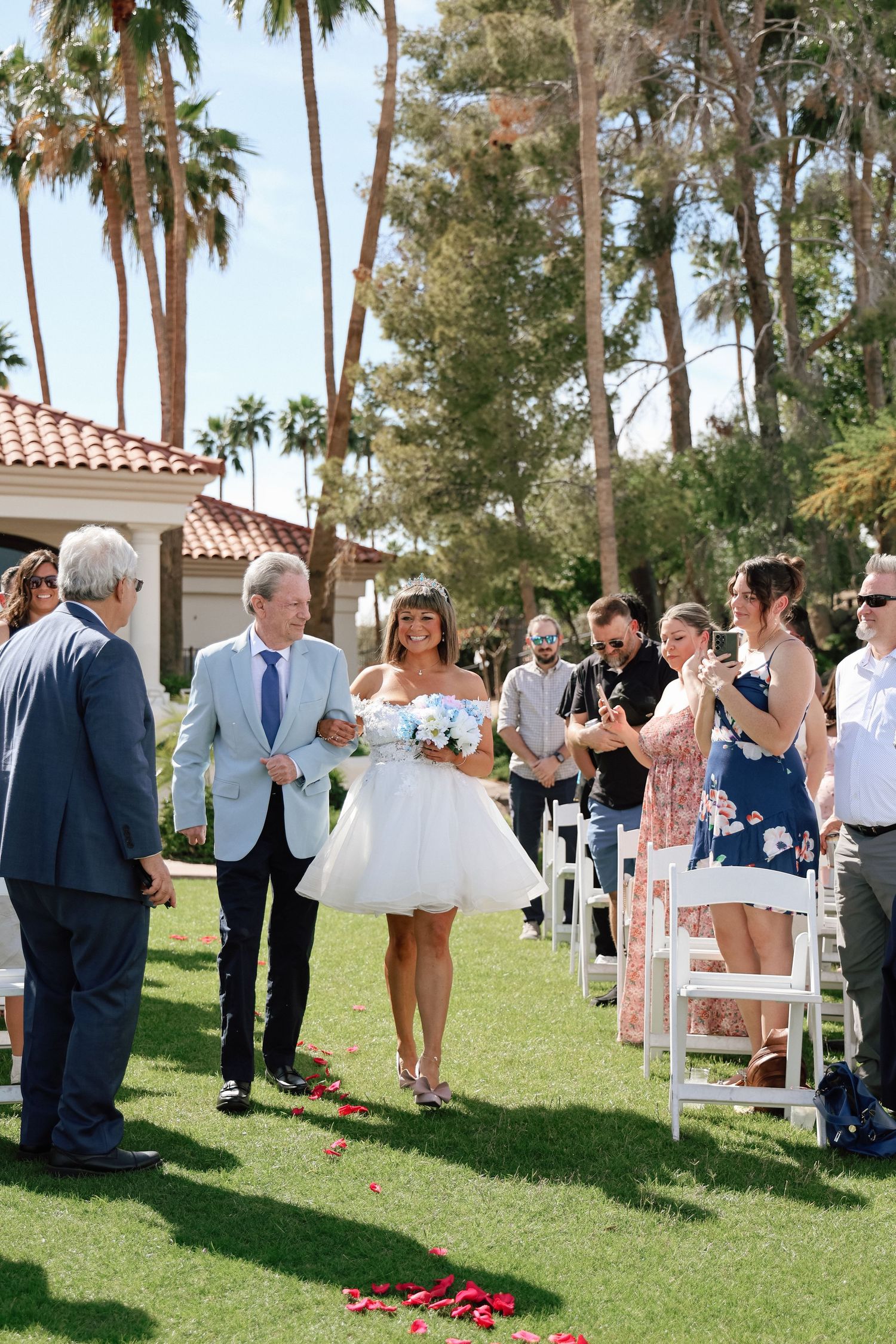 Wedding guests gather outdoors under palm trees on a lush green lawn during a sunny ceremony.