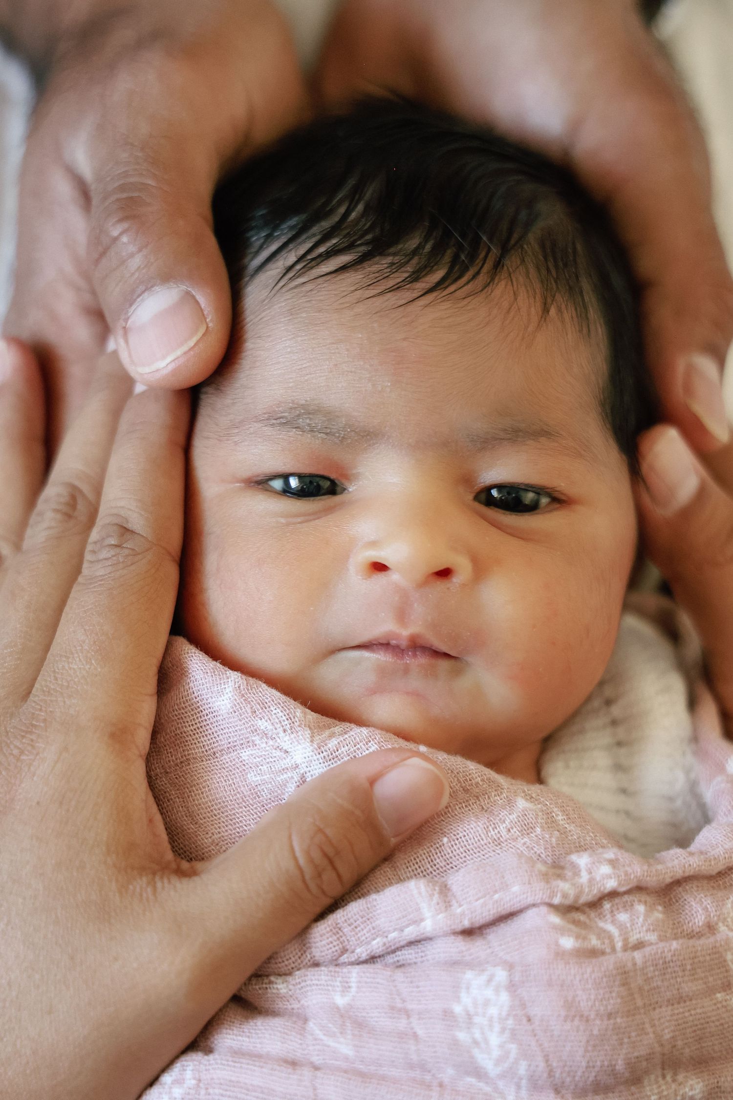 Newborn baby with dark hair cradled gently in adult hands, wrapped in a soft pink blanket, looking alert.