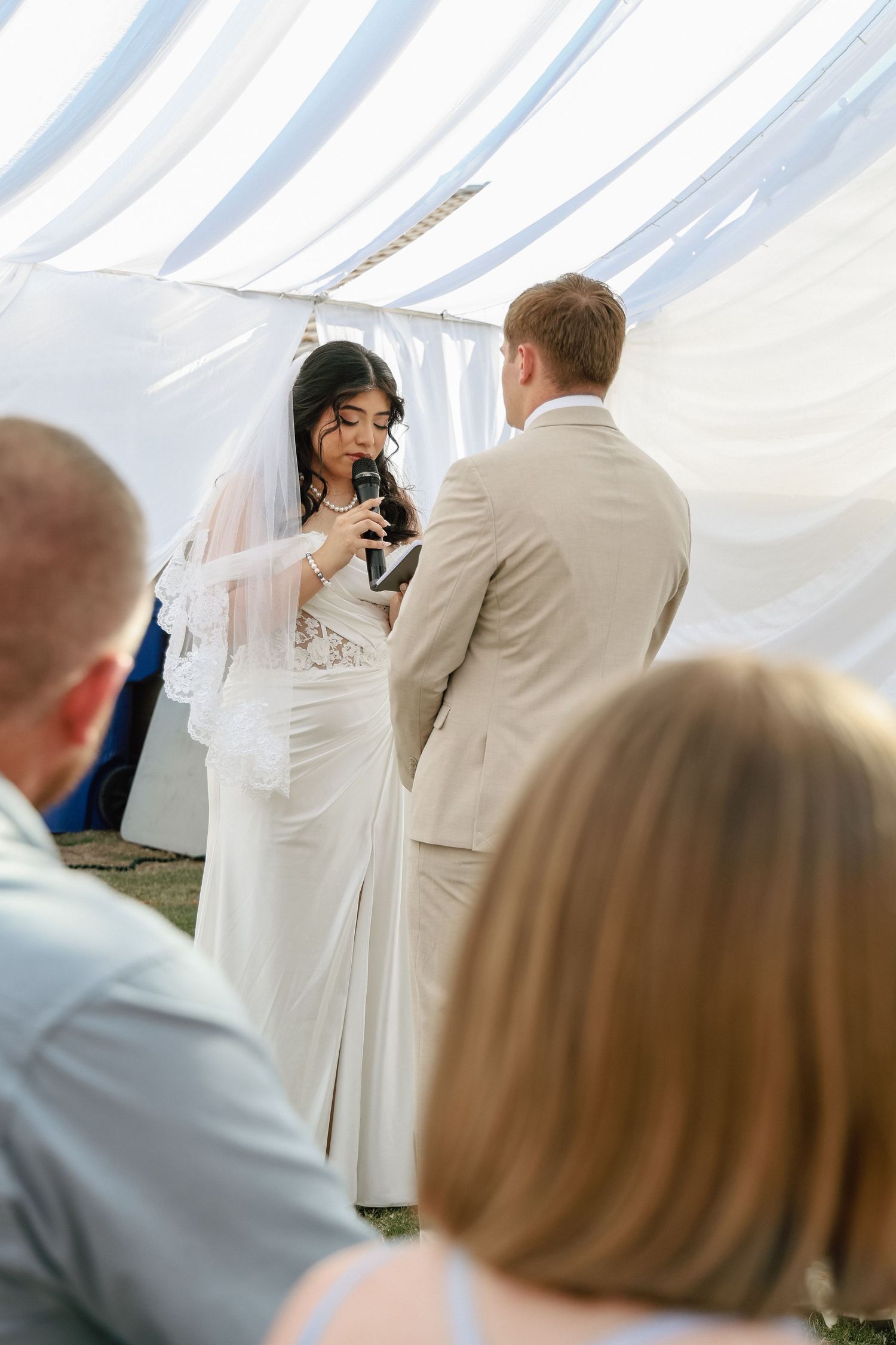 Bride in white dress and veil reads vows into microphone to groom in tan suit at outdoor wedding ceremony.