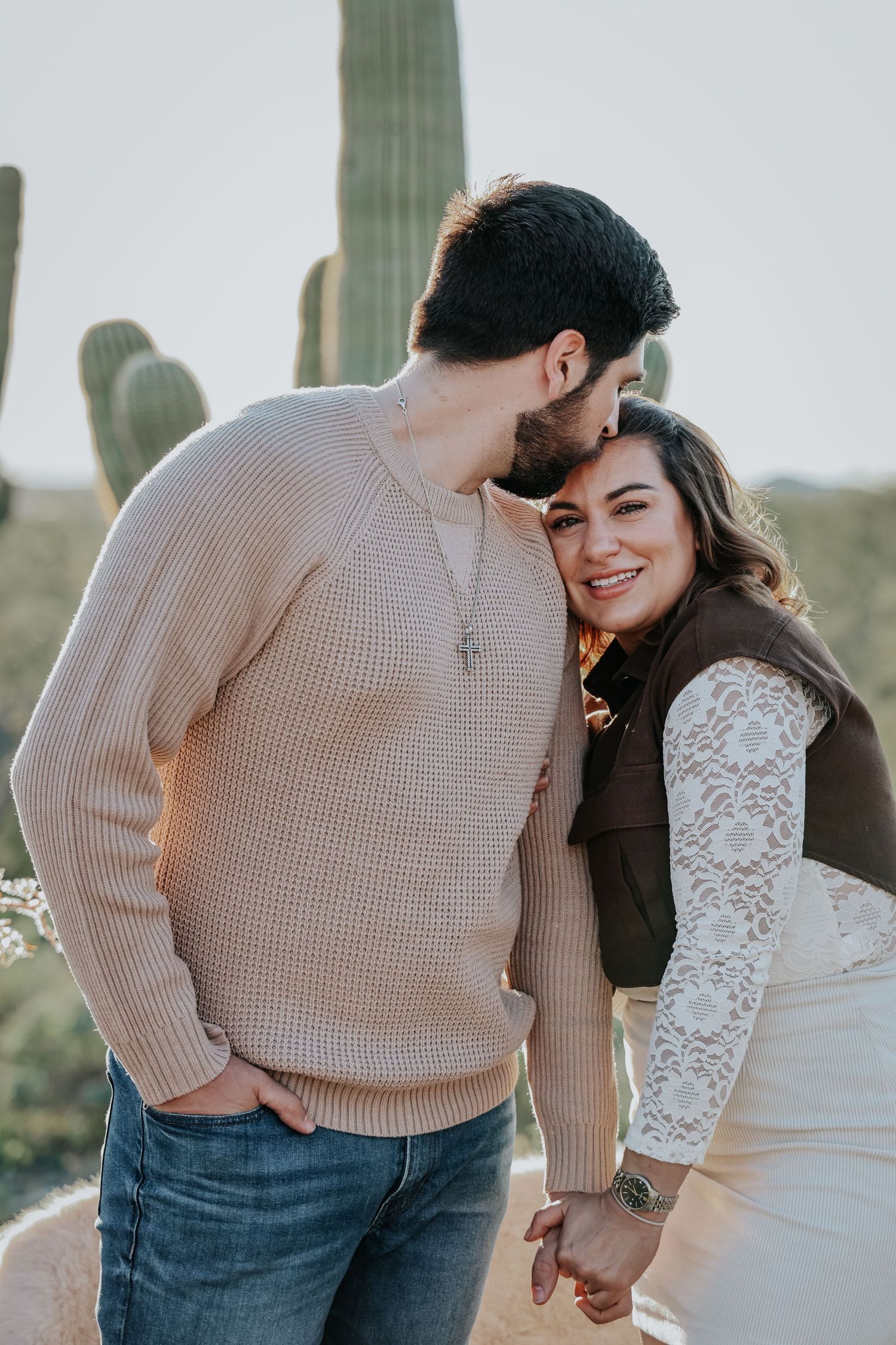 Couple sharing a romantic moment in front of saguaro cacti, woman smiling as man kisses her cheek.