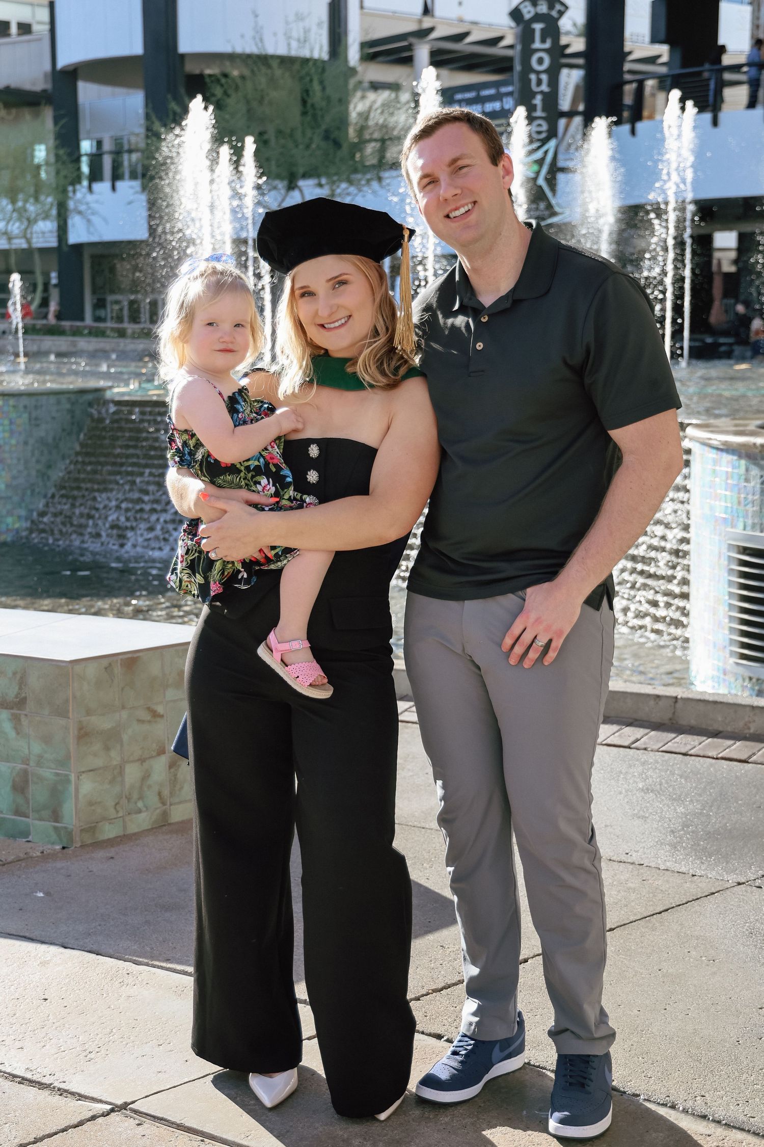 A woman in graduation cap holds a toddler beside a man, posing in front of a fountain outdoors.