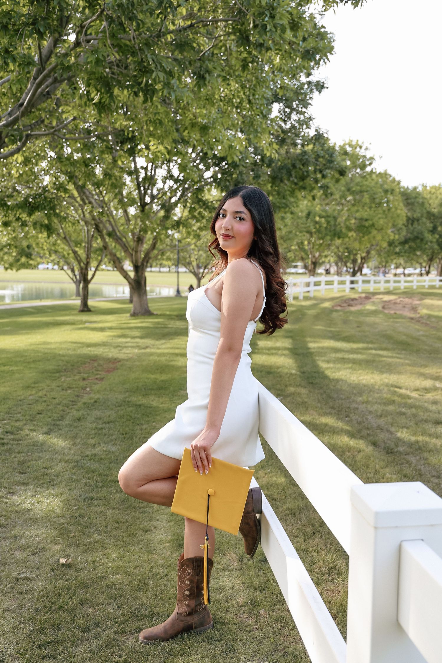 Young woman in white dress and cowboy boots holds graduation cap while leaning on white fence in park.