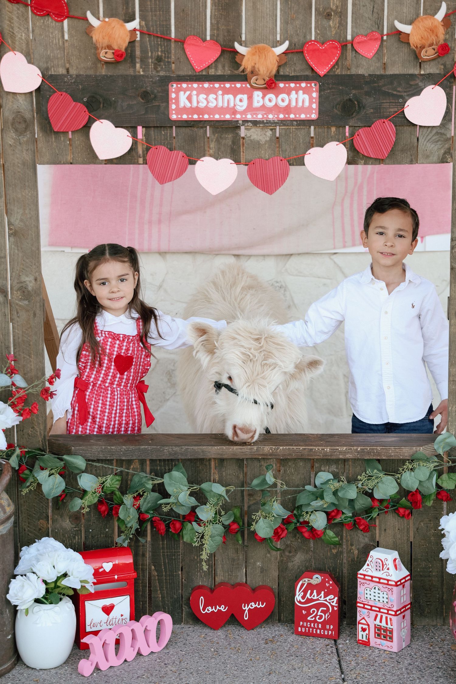 Two children pose at a Valentine's Day kissing booth with a fluffy white Highland calf, surrounded by heart decorations.
