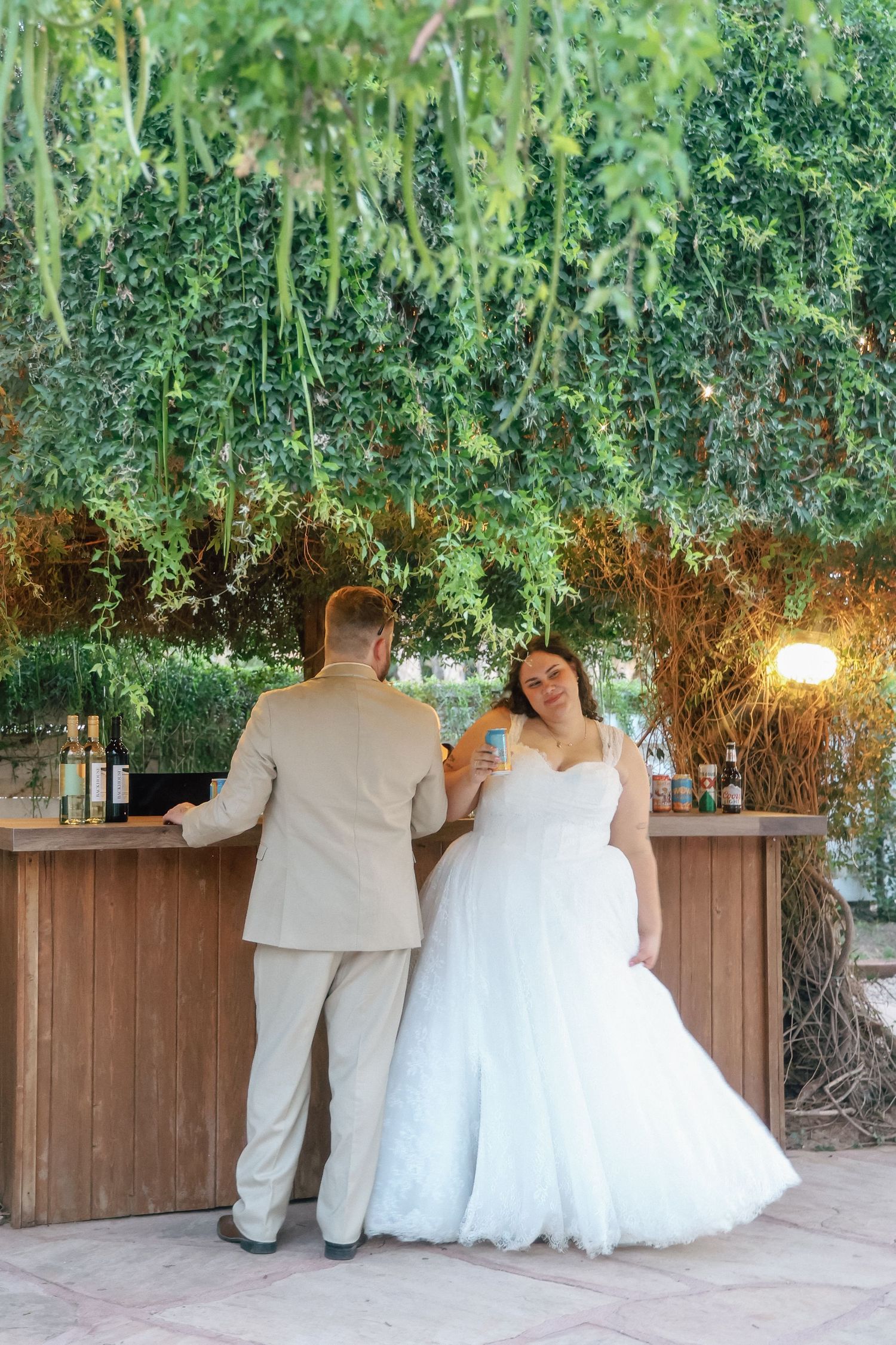 Bride in white gown and groom in tan suit stand at outdoor bar surrounded by lush green hanging foliage.