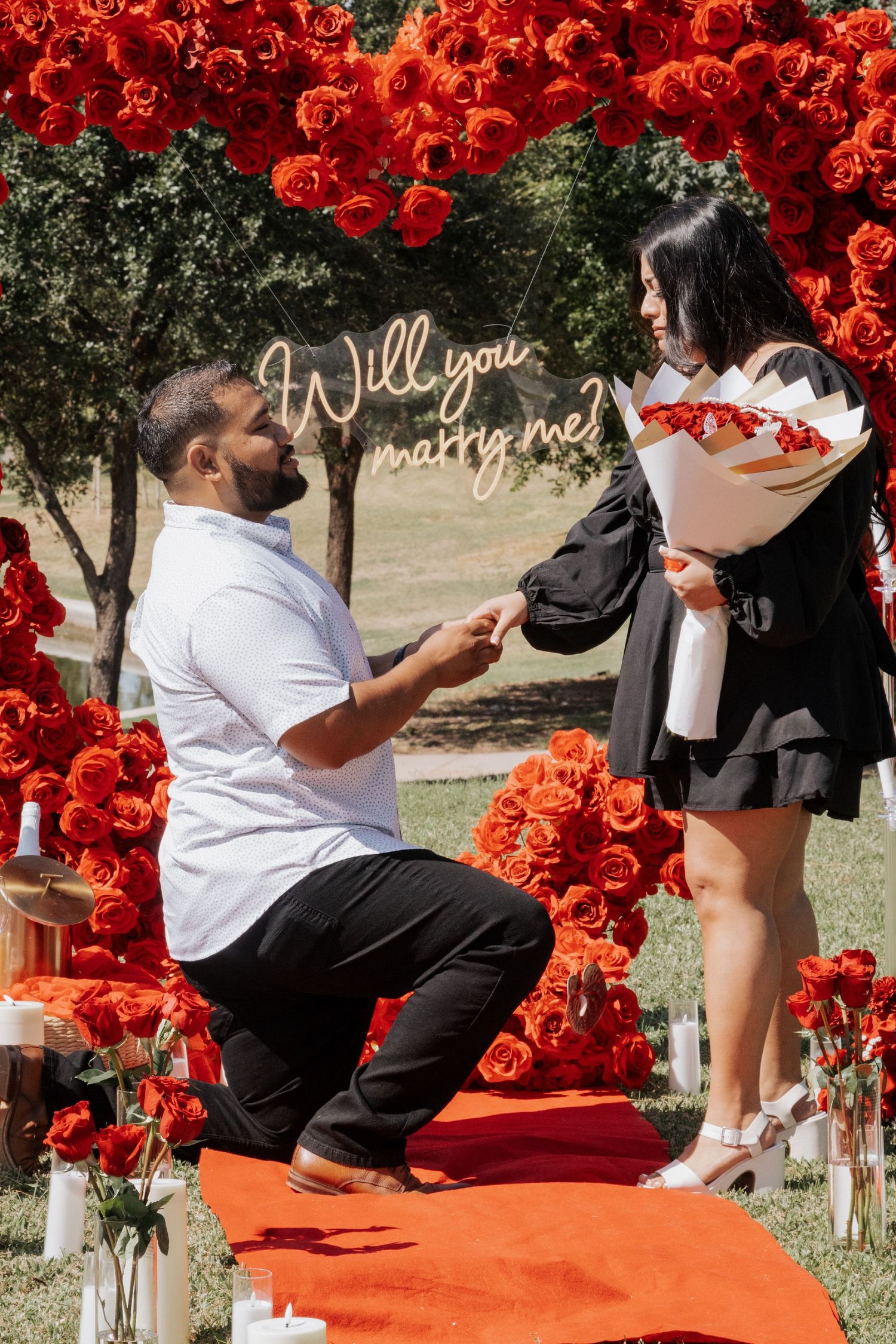 Man proposes on one knee to woman amid red roses arch with Will You Marry Me neon sign outdoors.
