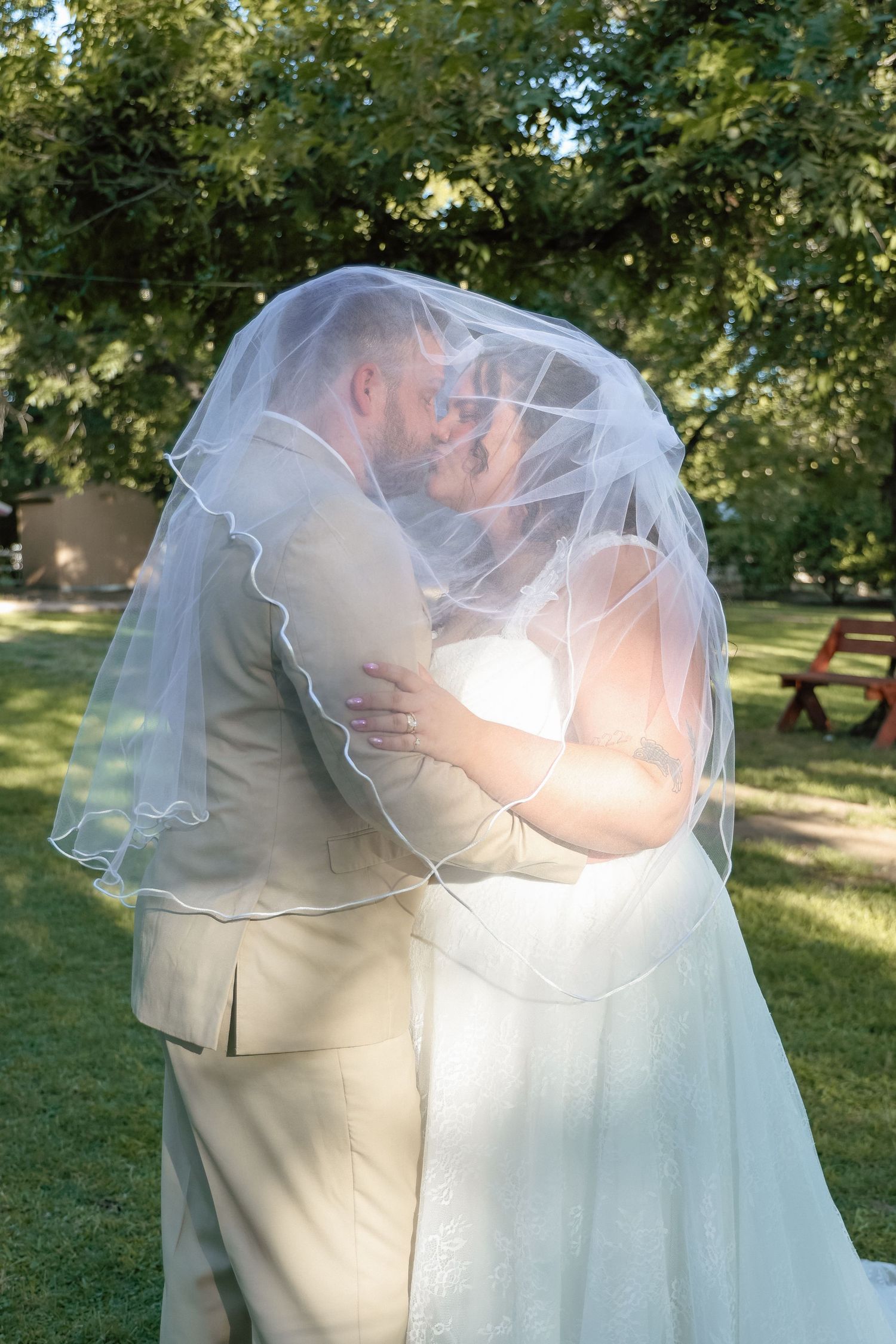 Bride and groom sharing a kiss under a flowing white veil in a lush green outdoor garden setting.