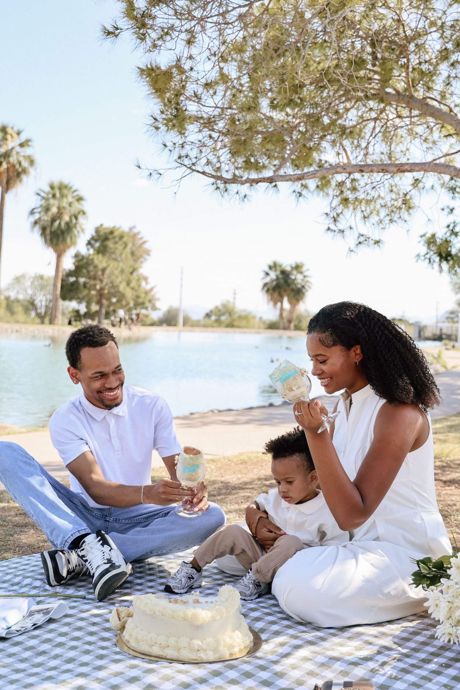 Happy Black family enjoying a birthday cake picnic by the lake in a sunny park setting.