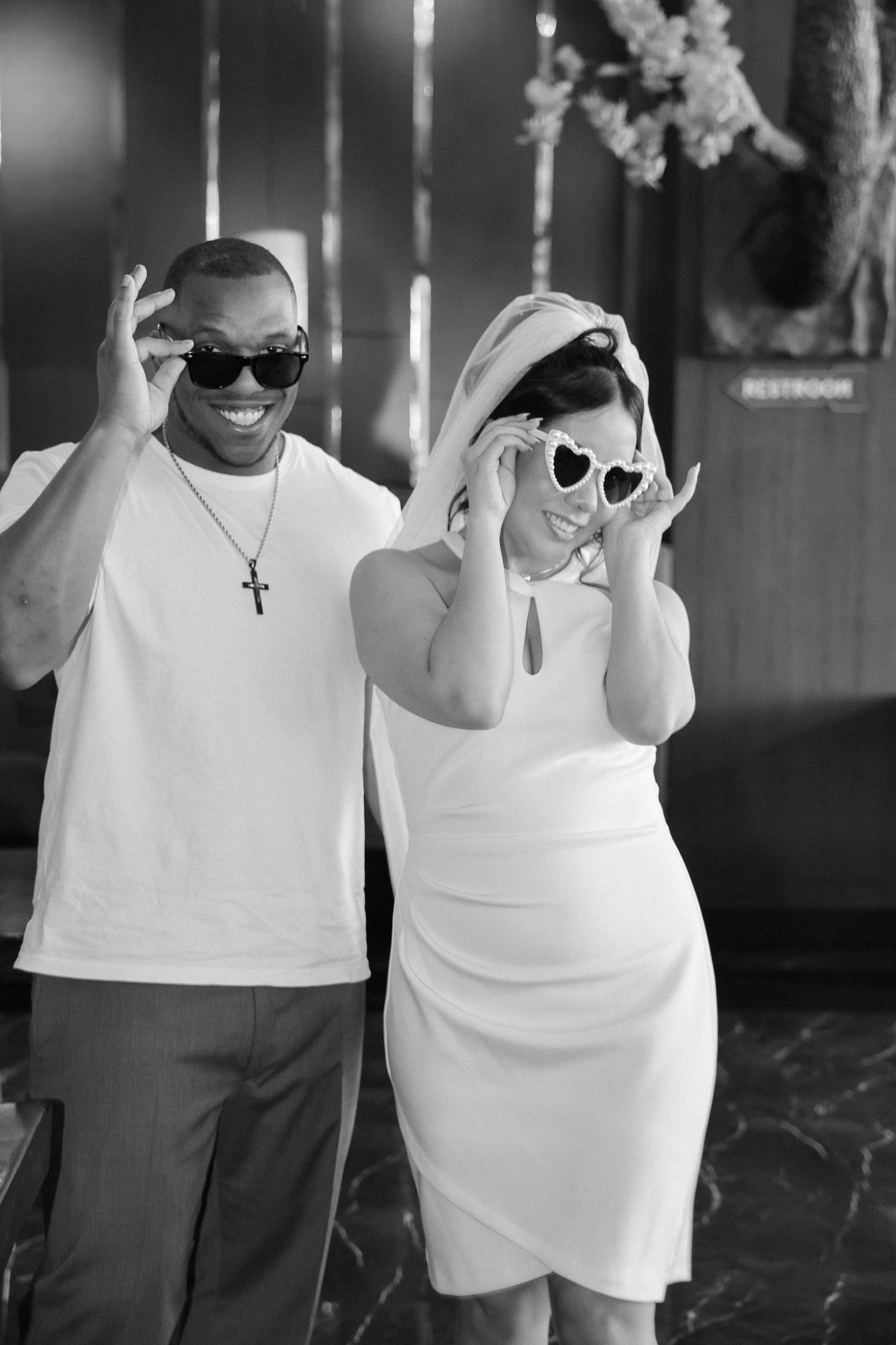 A couple wearing white and grey outfits pose playfully with sunglasses in a black and white photo series.