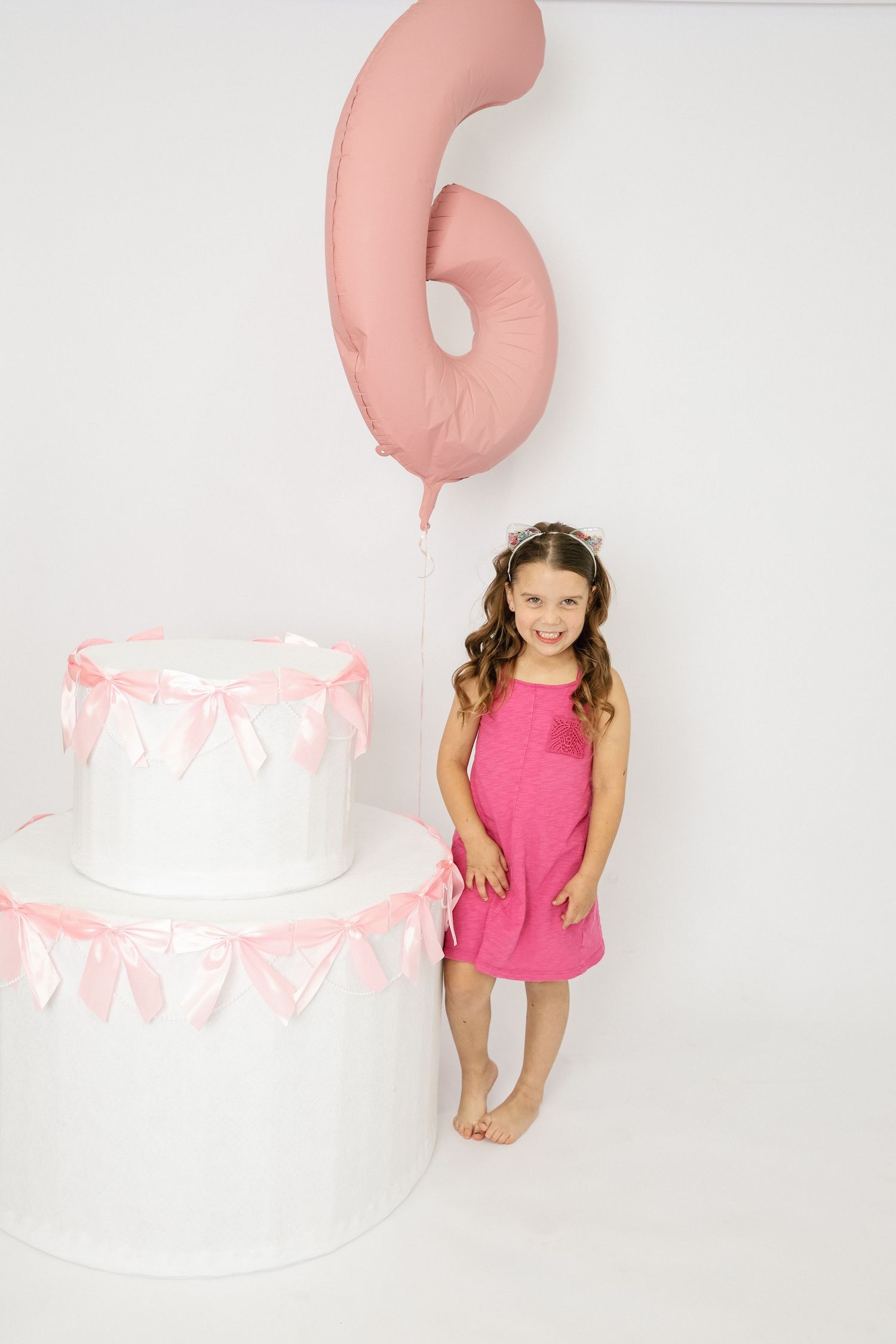 A smiling girl in a pink dress stands next to a white birthday cake with a pink number 6 balloon.