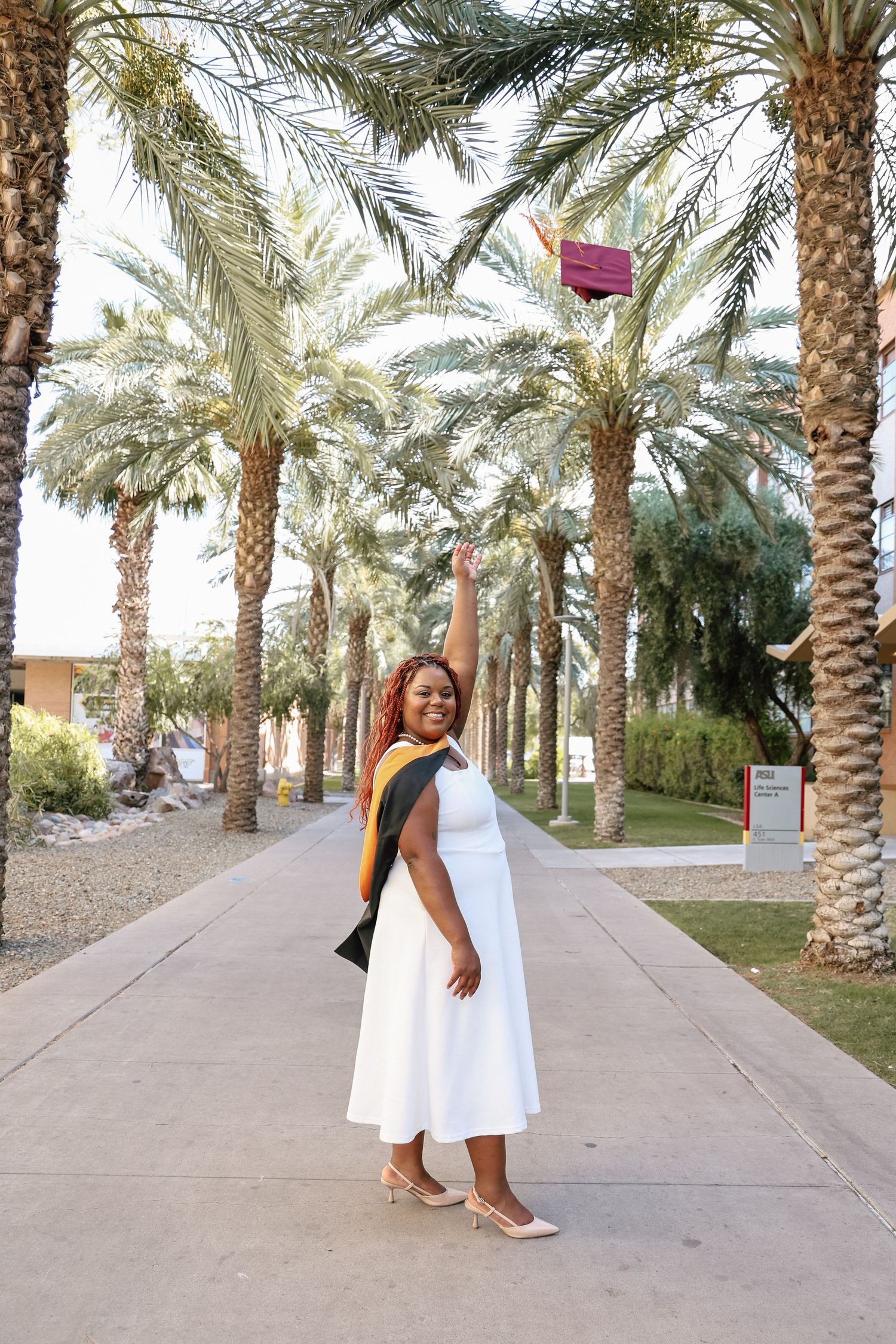 Woman in white dress and graduation sash tosses cap among palm trees, celebrating academic achievement outdoors.