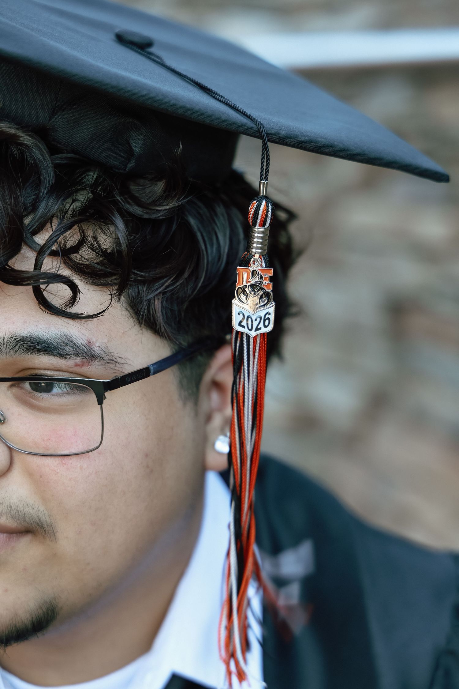 Young man in black graduation cap and gown with 2026 tassel, wearing glasses, celebrating academic milestone.