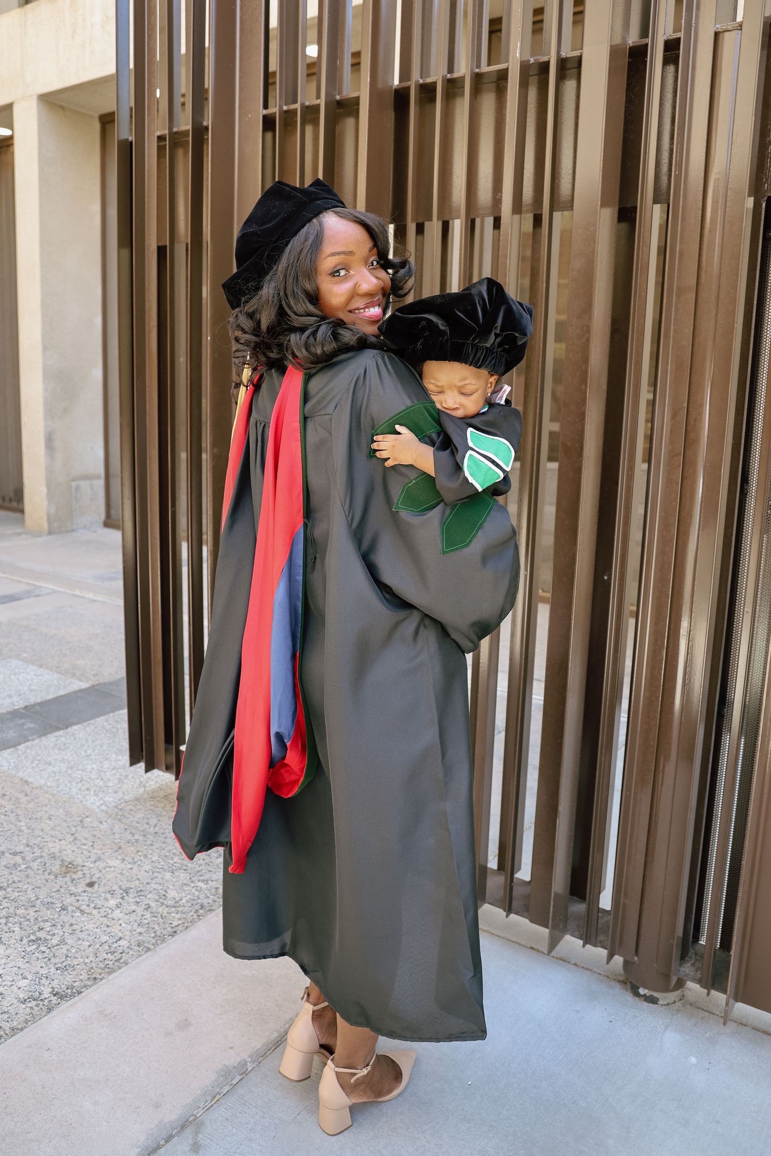 Smiling graduate in black cap and gown holds sleeping baby dressed in matching graduation attire outdoors.
