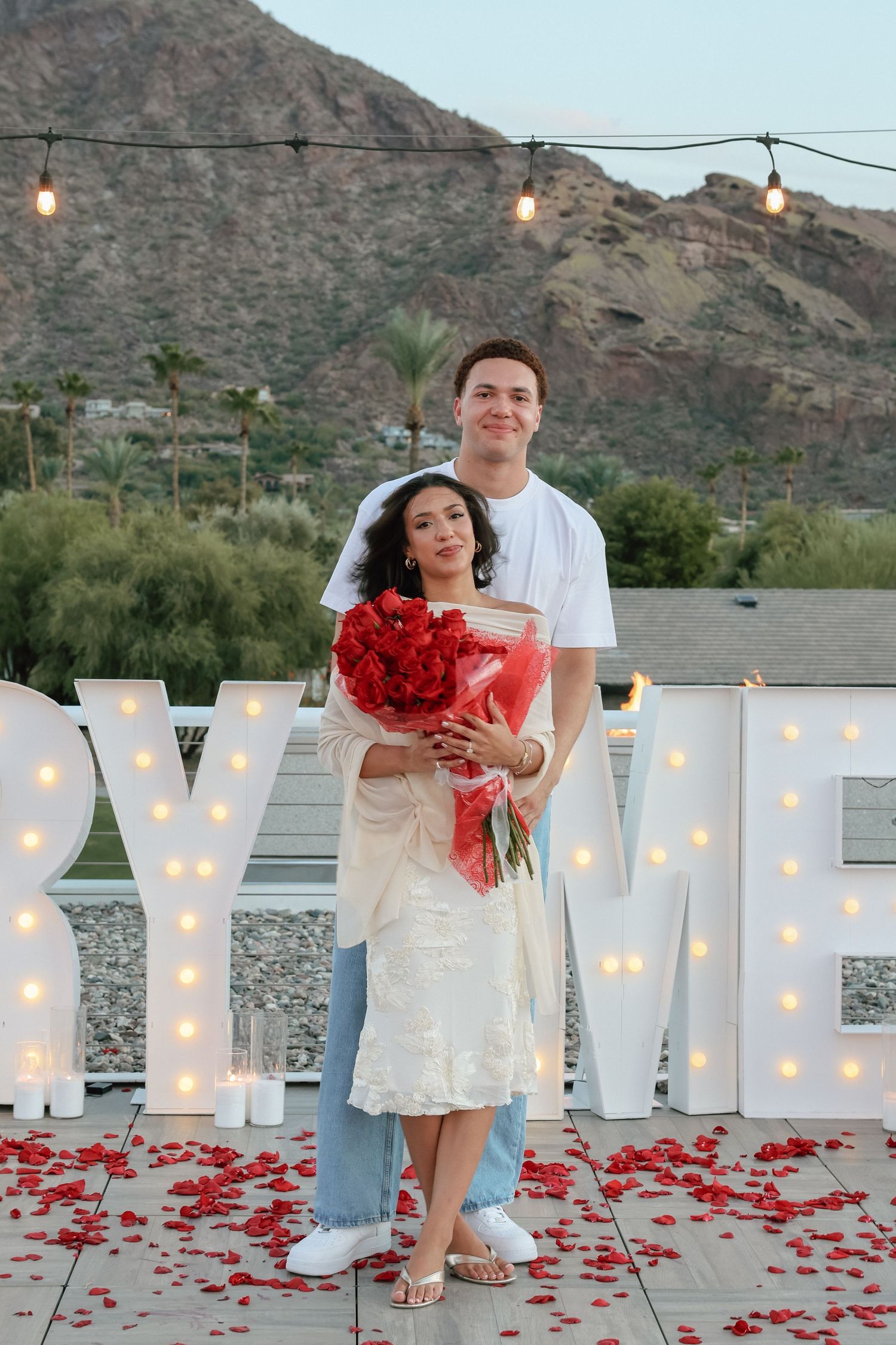 Couple poses with red roses bouquet in front of illuminated marry me letters and rose petals with mountain backdrop.