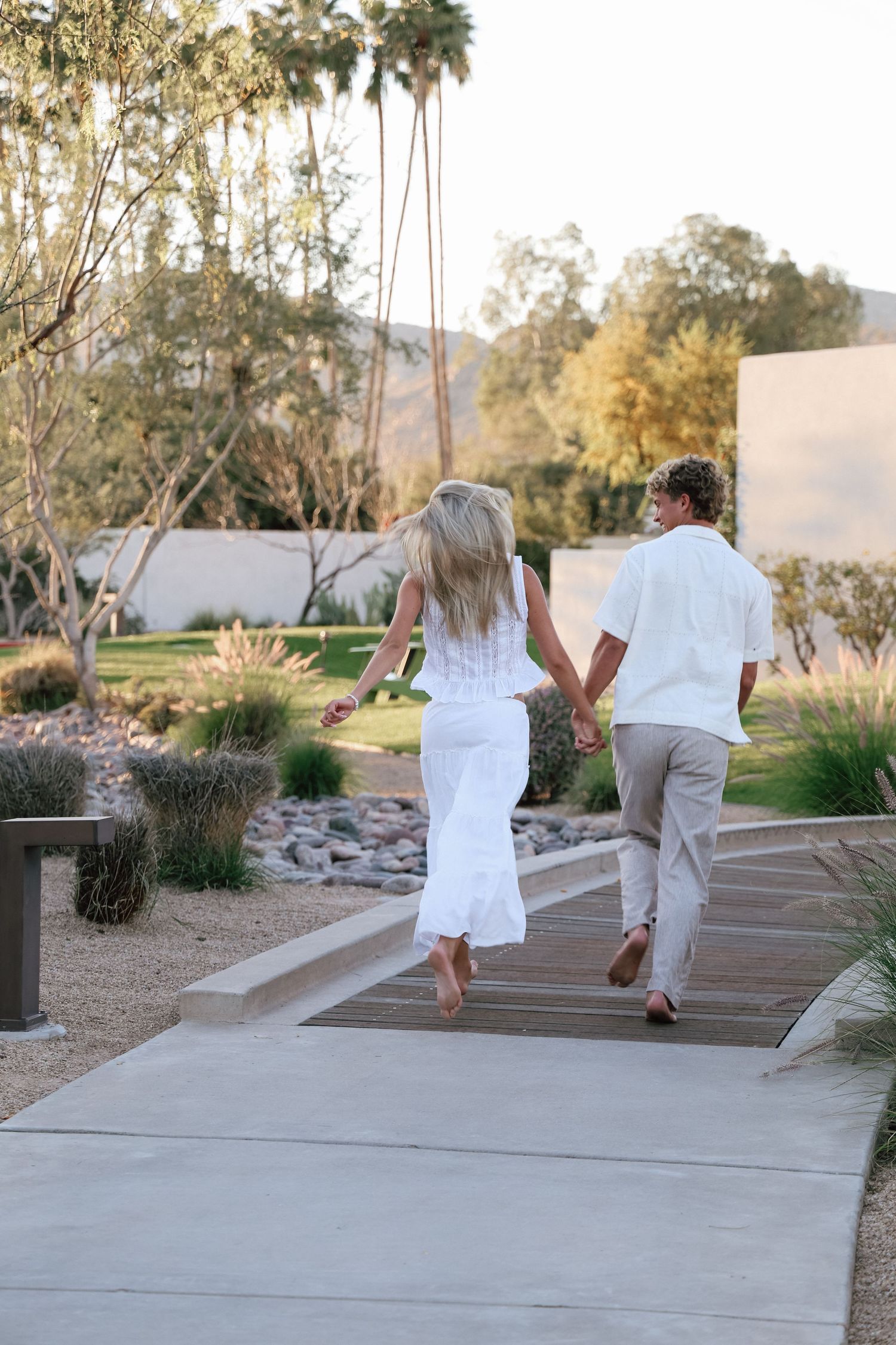 Couple in white outfits holding hands walking along a sunny resort pathway surrounded by desert landscaping.