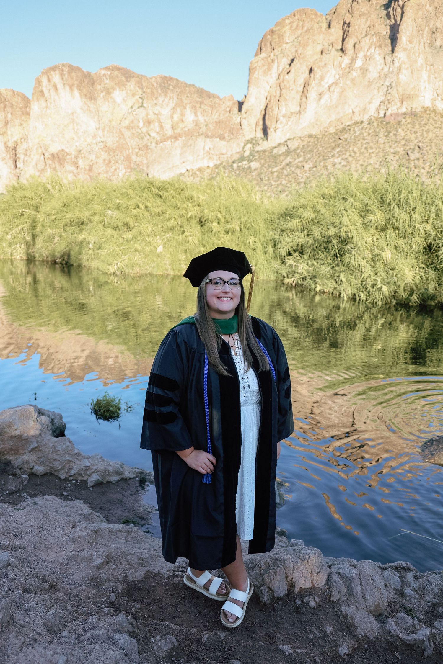 Woman in black doctoral graduation regalia standing on rocks beside a reflective river with scenic mountain backdrop.