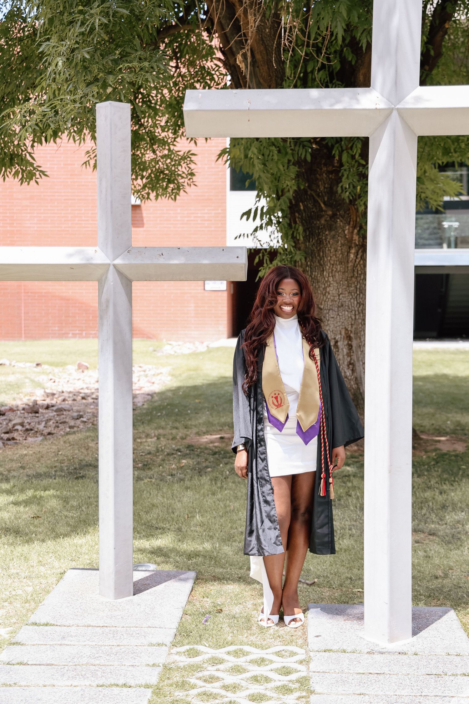 Young woman in graduation cap and gown with stole poses proudly between white cross sculptures on a sunny campus.