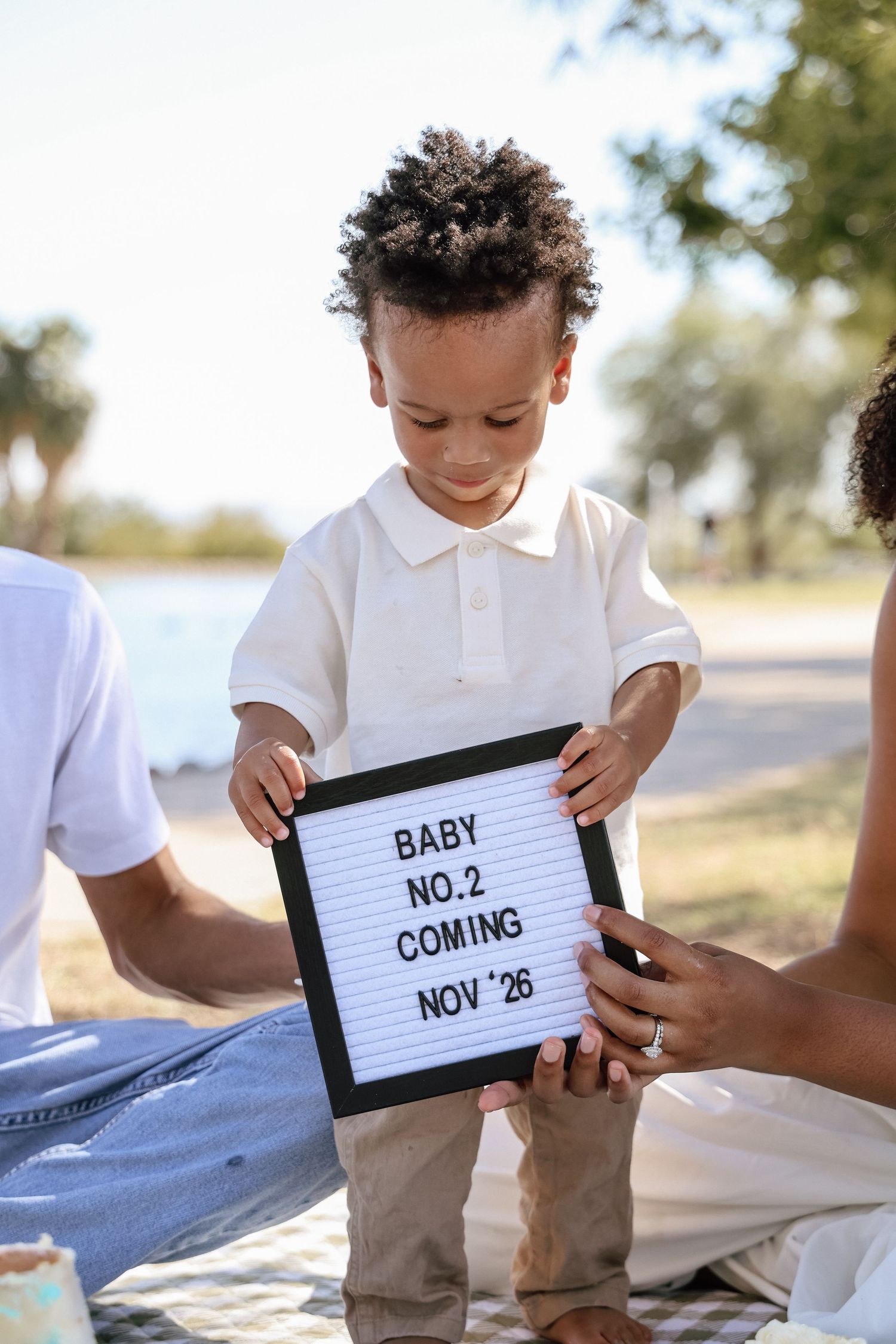 A young child holds a letter board announcing Baby No. 2 coming Nov 26 during an outdoor family photo shoot.