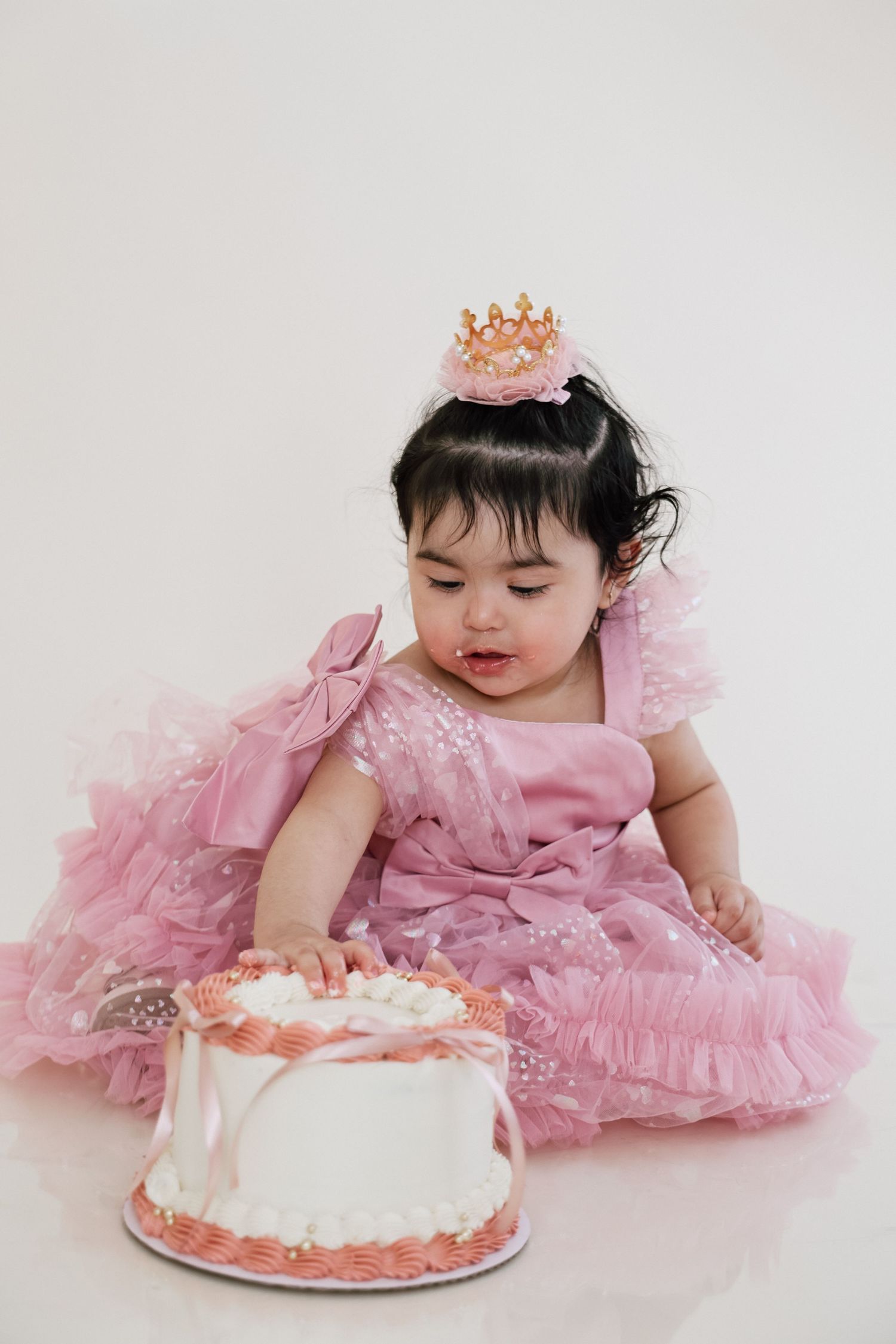 Baby in pink tulle dress lying on floor reaching for white birthday cake in elegant first birthday photo session.