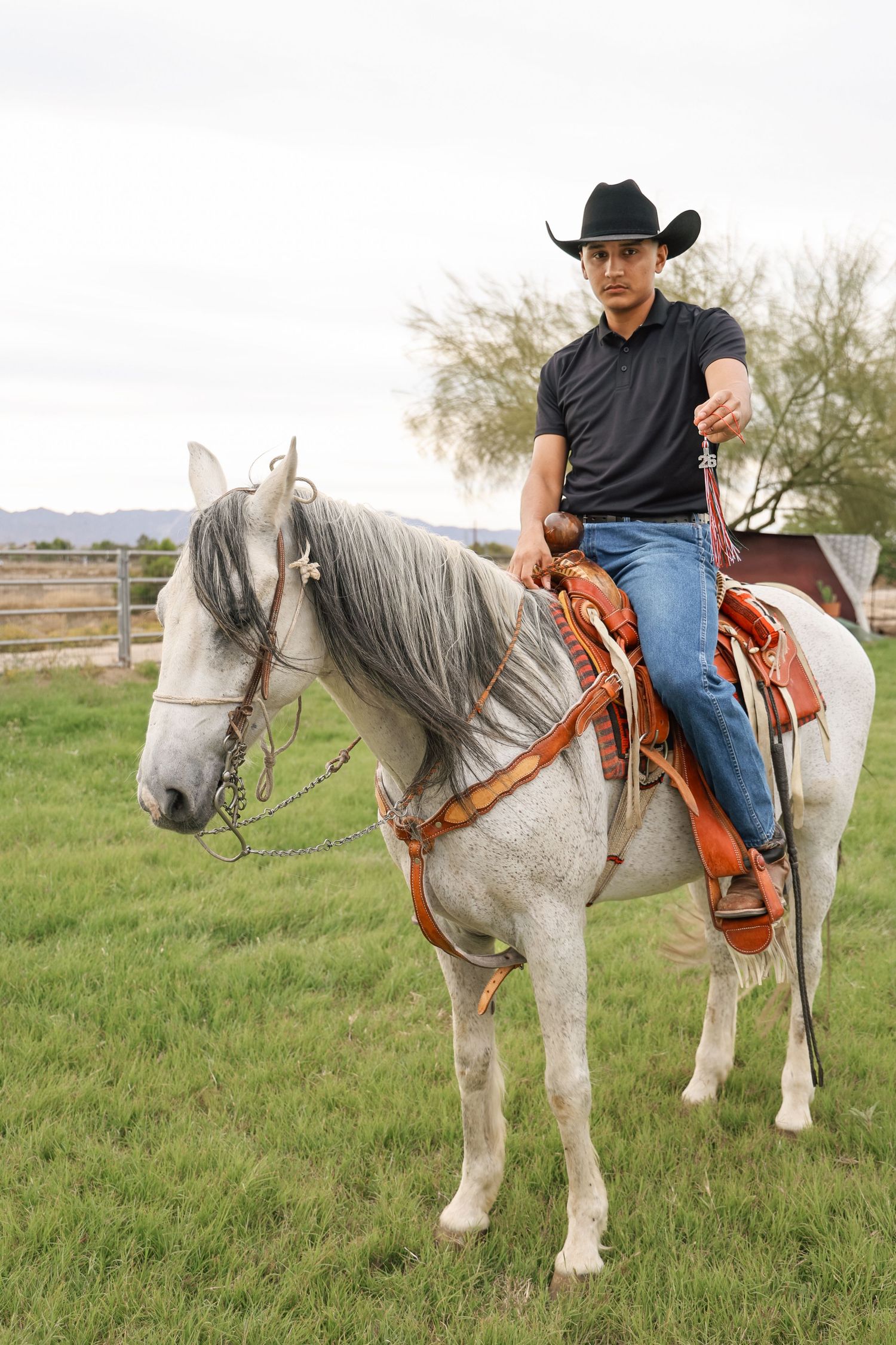 A young cowboy in a black hat and jeans rides a gray horse in a green pasture with a Western saddle.