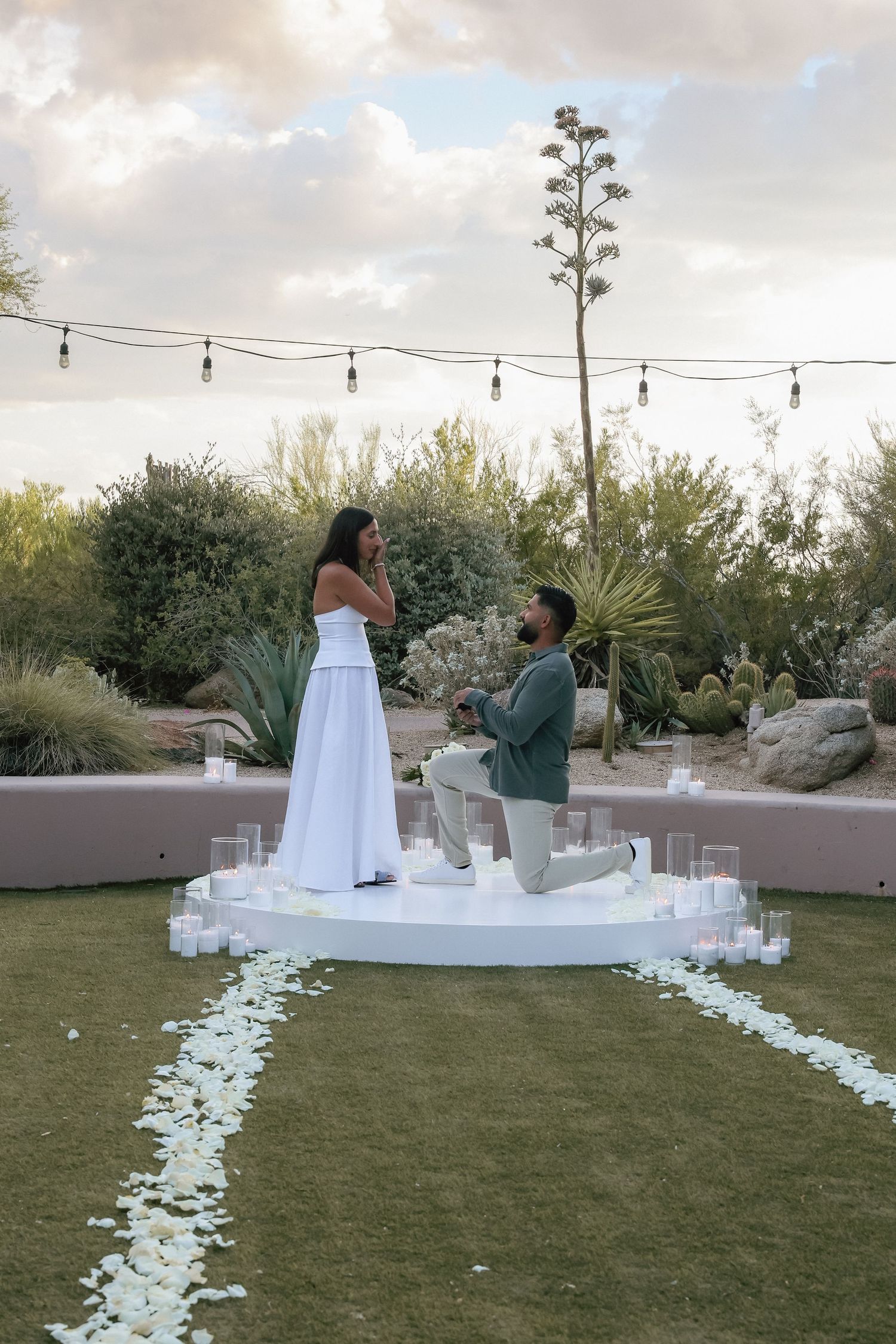 Man proposes on white platform surrounded by candles and rose petals in desert garden at sunset.