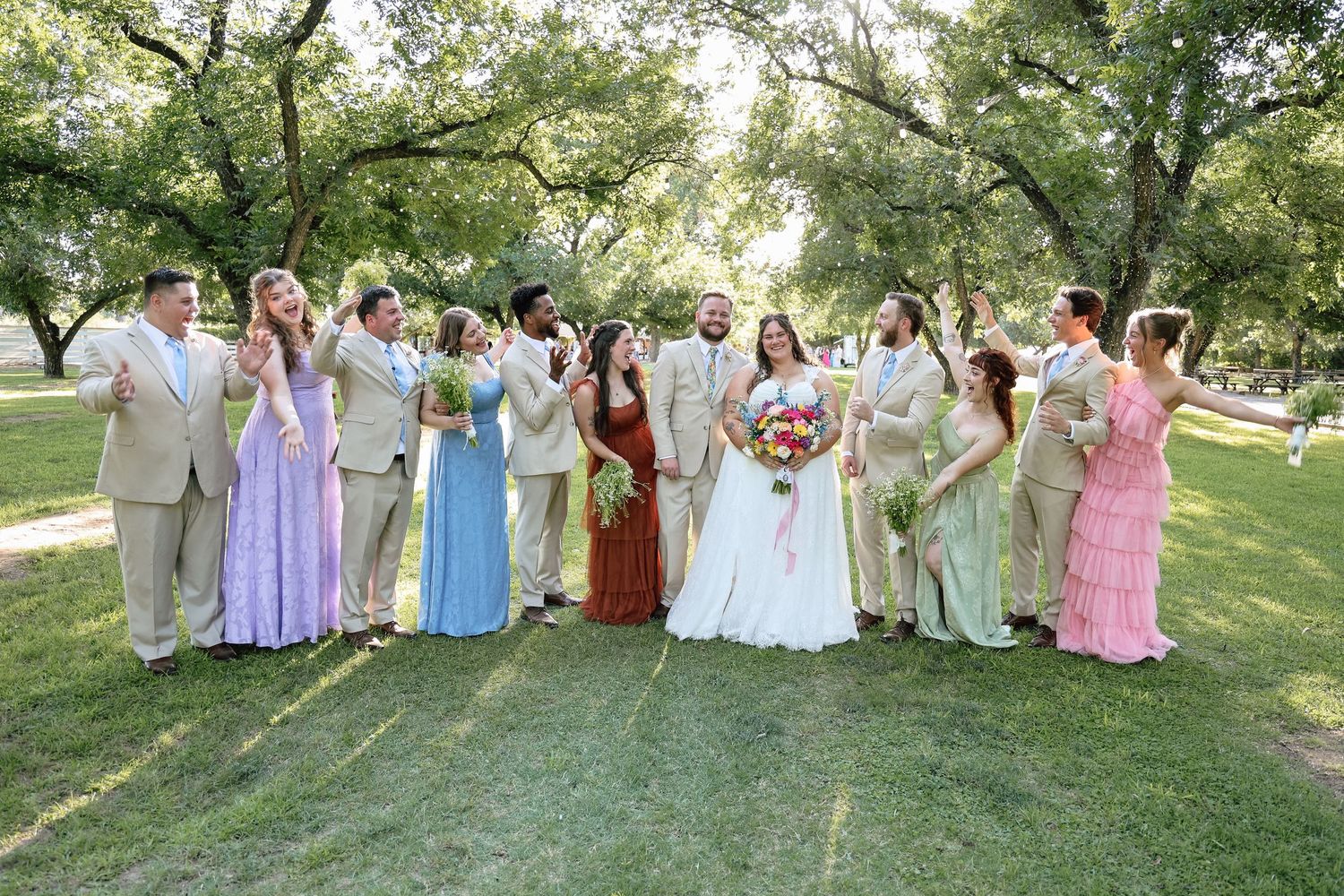 Wedding party in colorful gowns and tan suits celebrating outdoors under lush green trees on a sunny day.