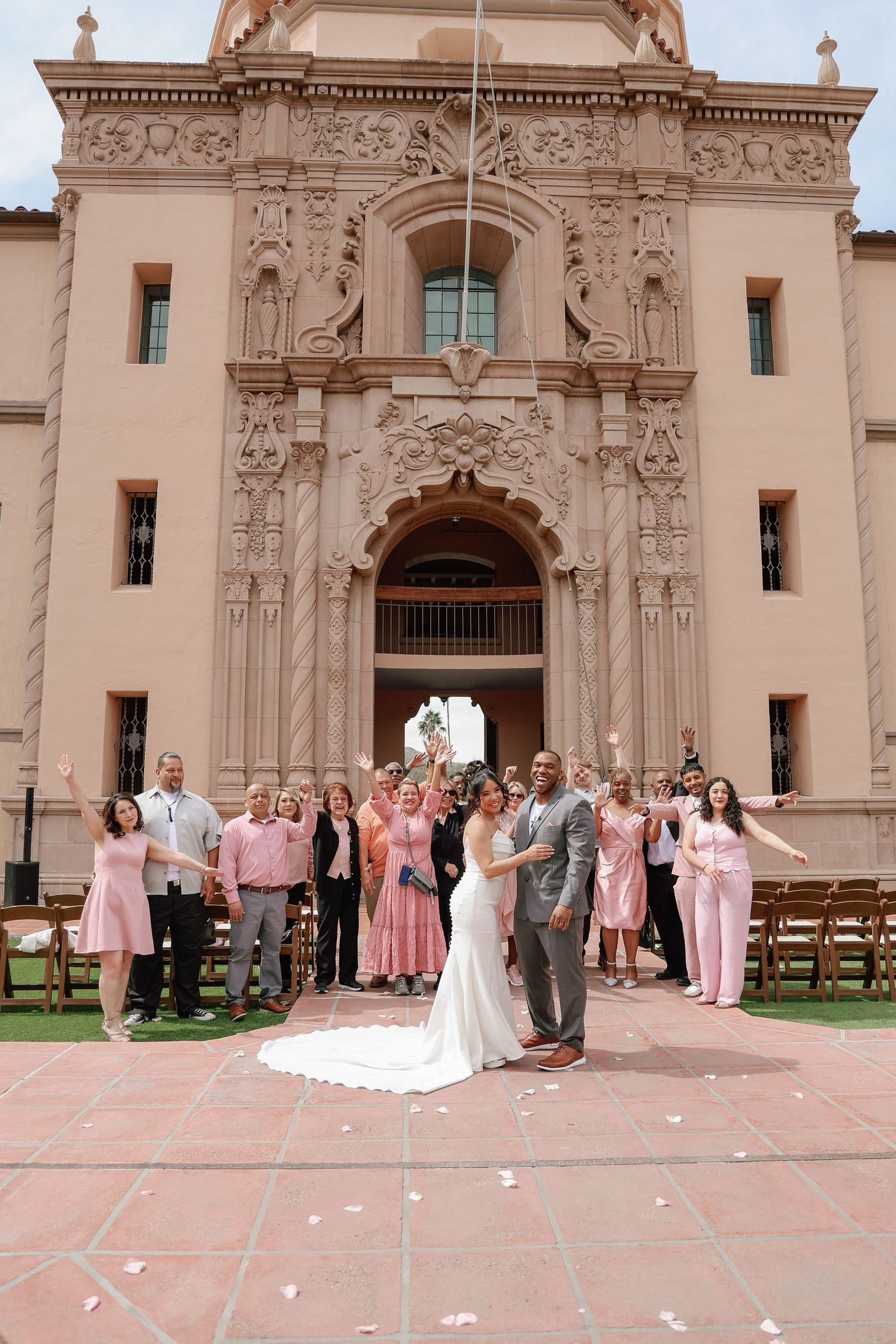 Bride and groom kiss before ornate Balboa Park building with cheering wedding guests in pink attire.