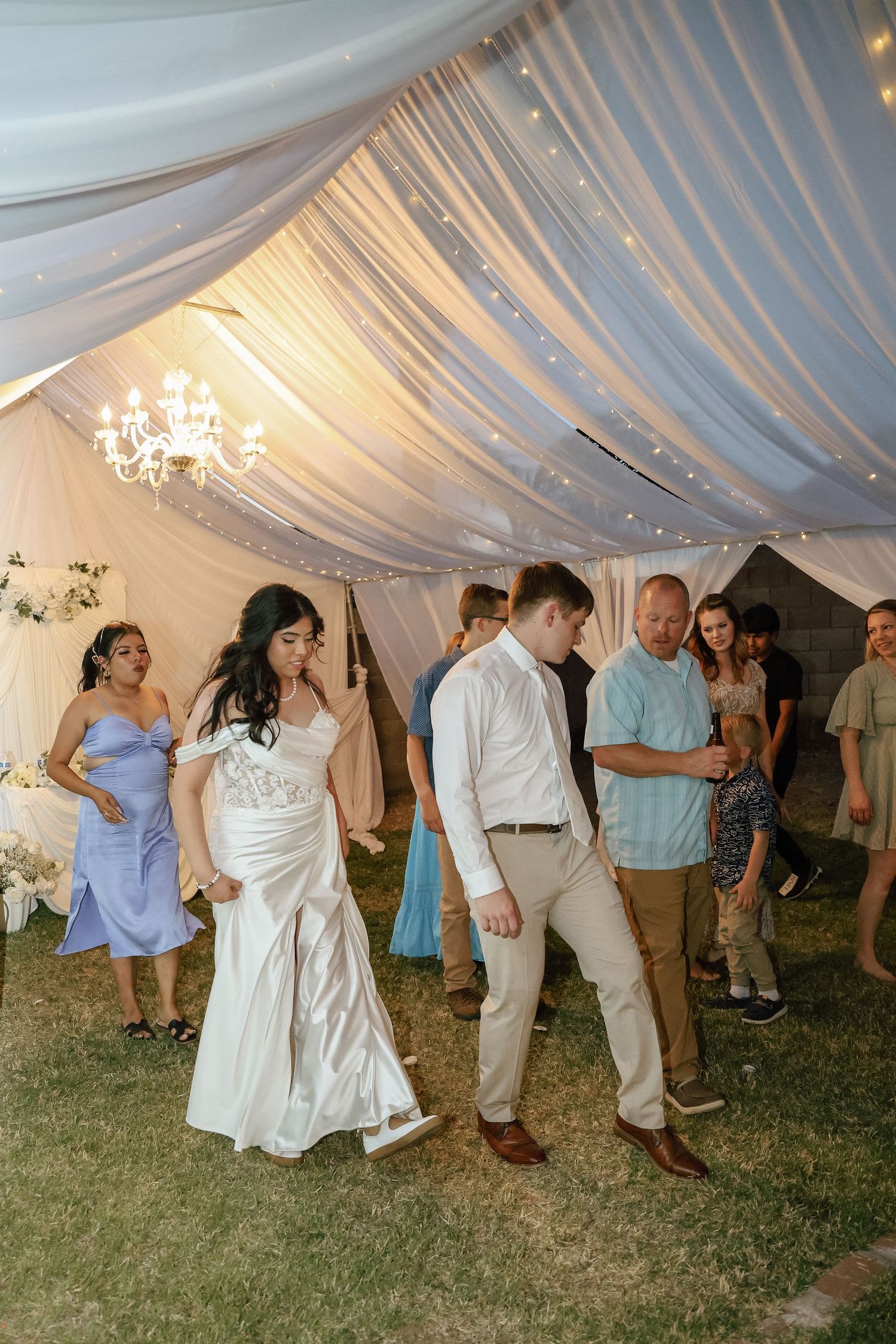 Bride and groom dance together at outdoor wedding reception under elegant draped tent with chandelier lighting.