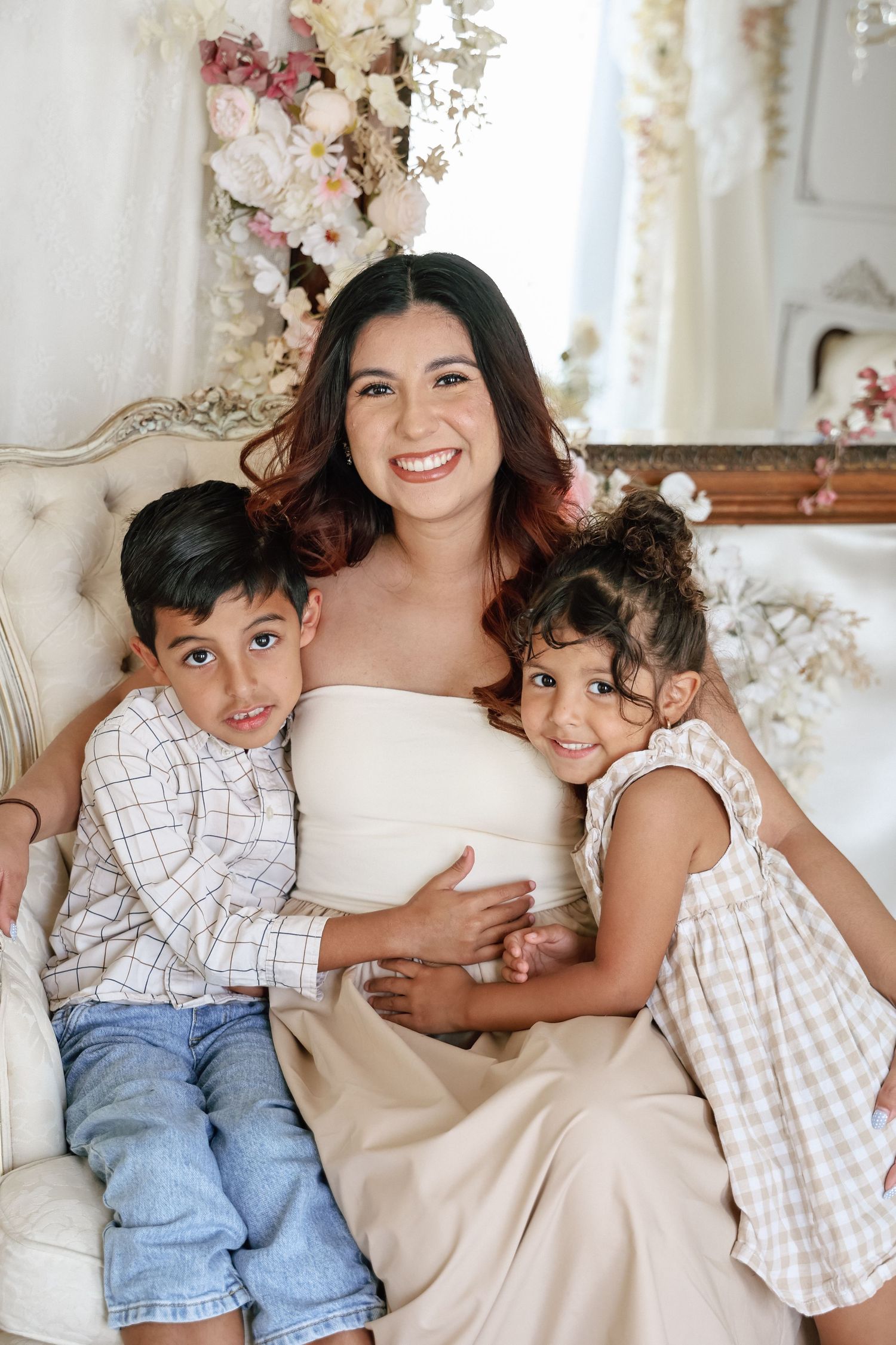 Pregnant woman in white dress smiling with two young children on elegant sofa with floral backdrop.